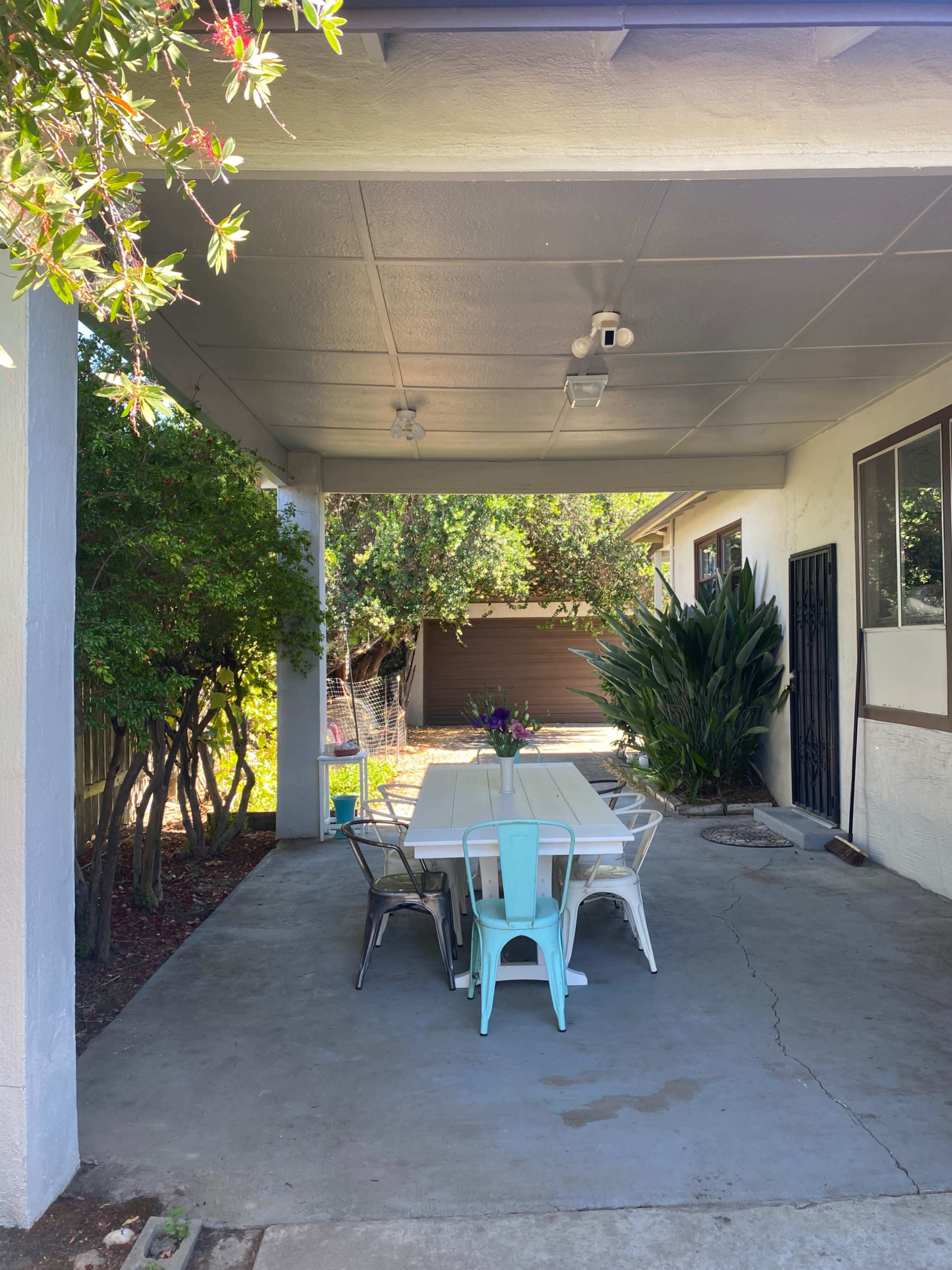 A shaded outdoor patio features a white table surrounded by metal and vintage-style chairs, with green foliage in the background.