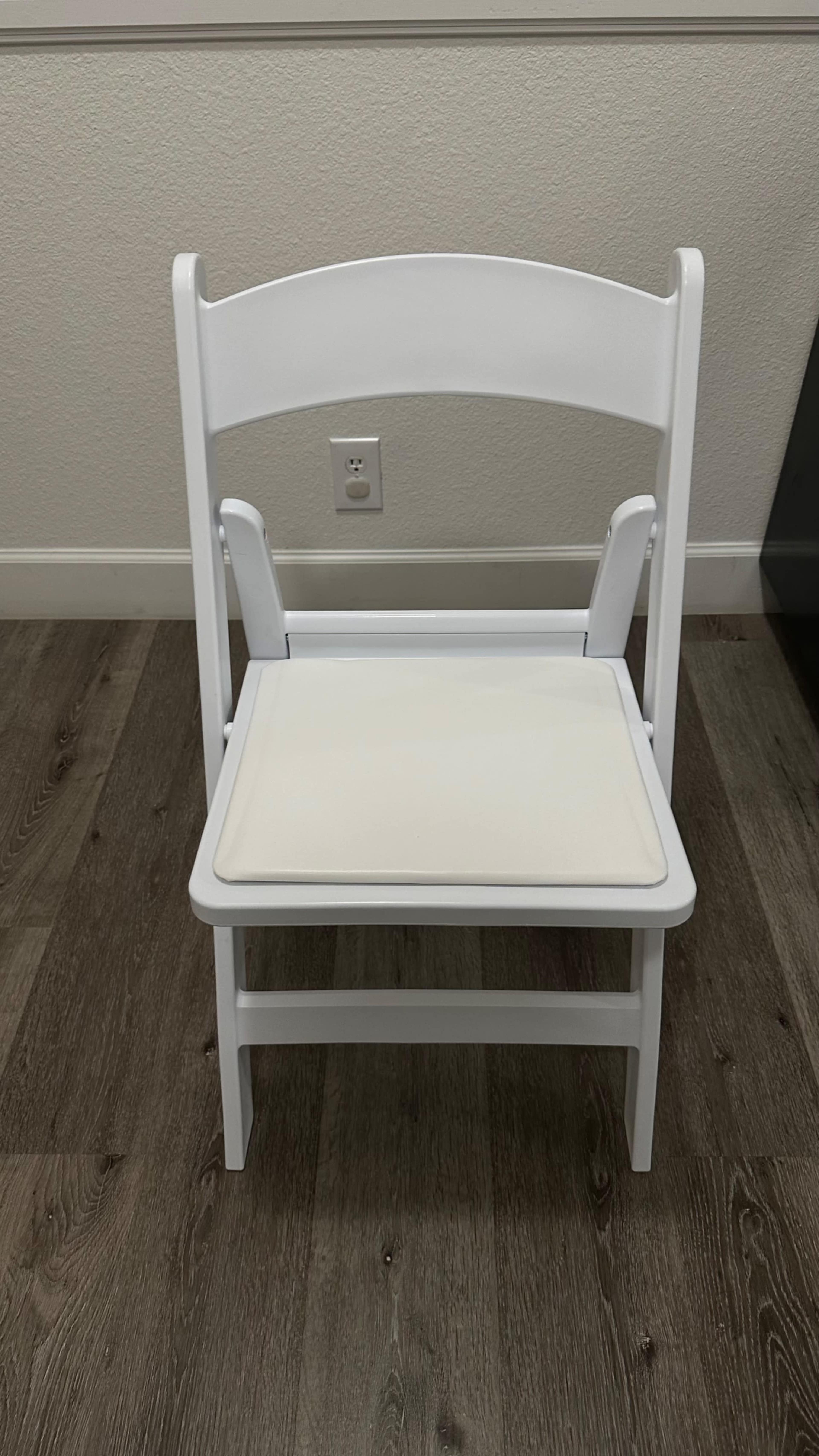 The image shows a white folding chair with a cushioned seat, placed on a wooden floor in a well-lit room.