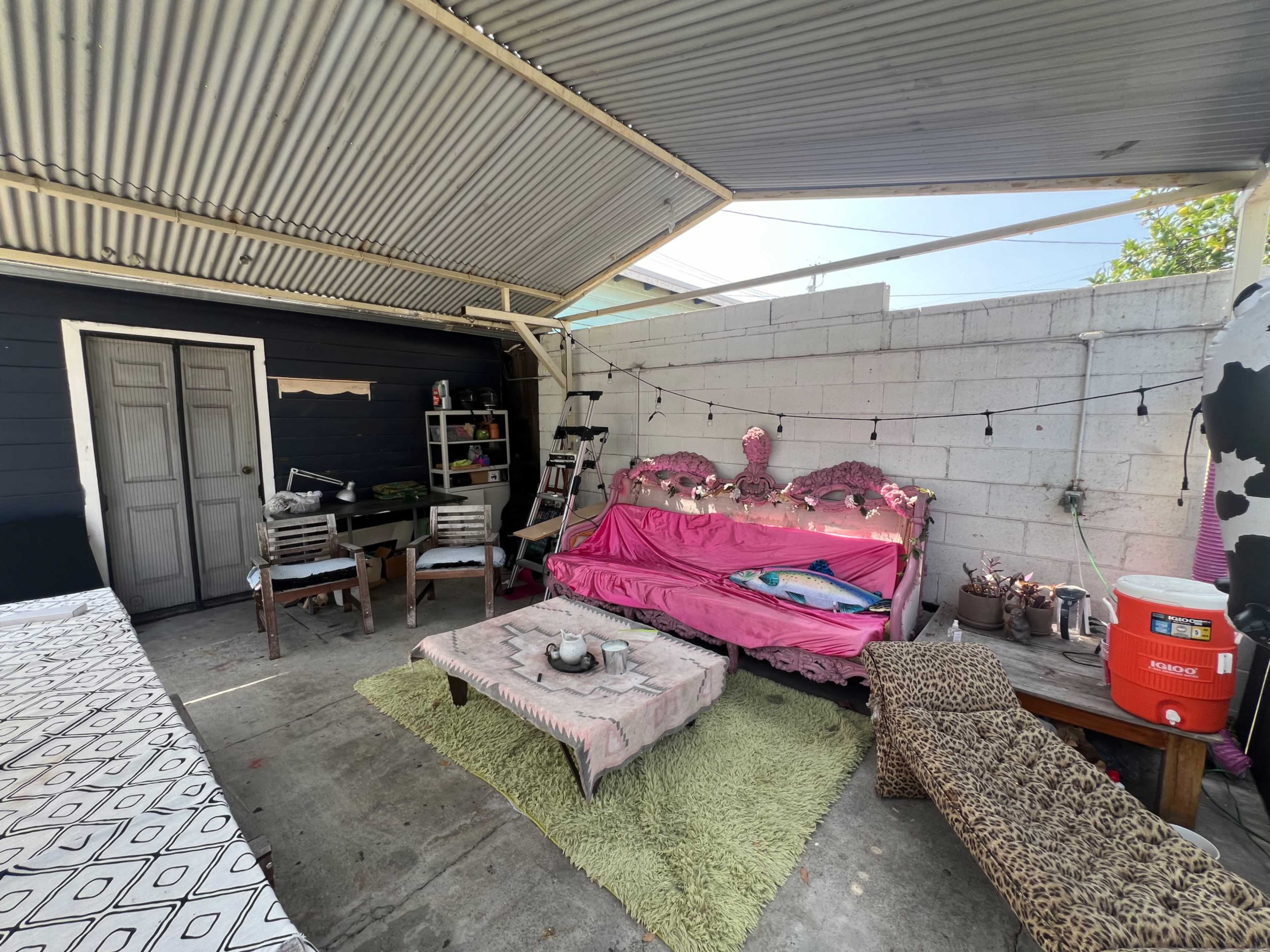 The image shows a shaded outdoor area with a pink sofa, wooden table, and various seating options, surrounded by a concrete wall.