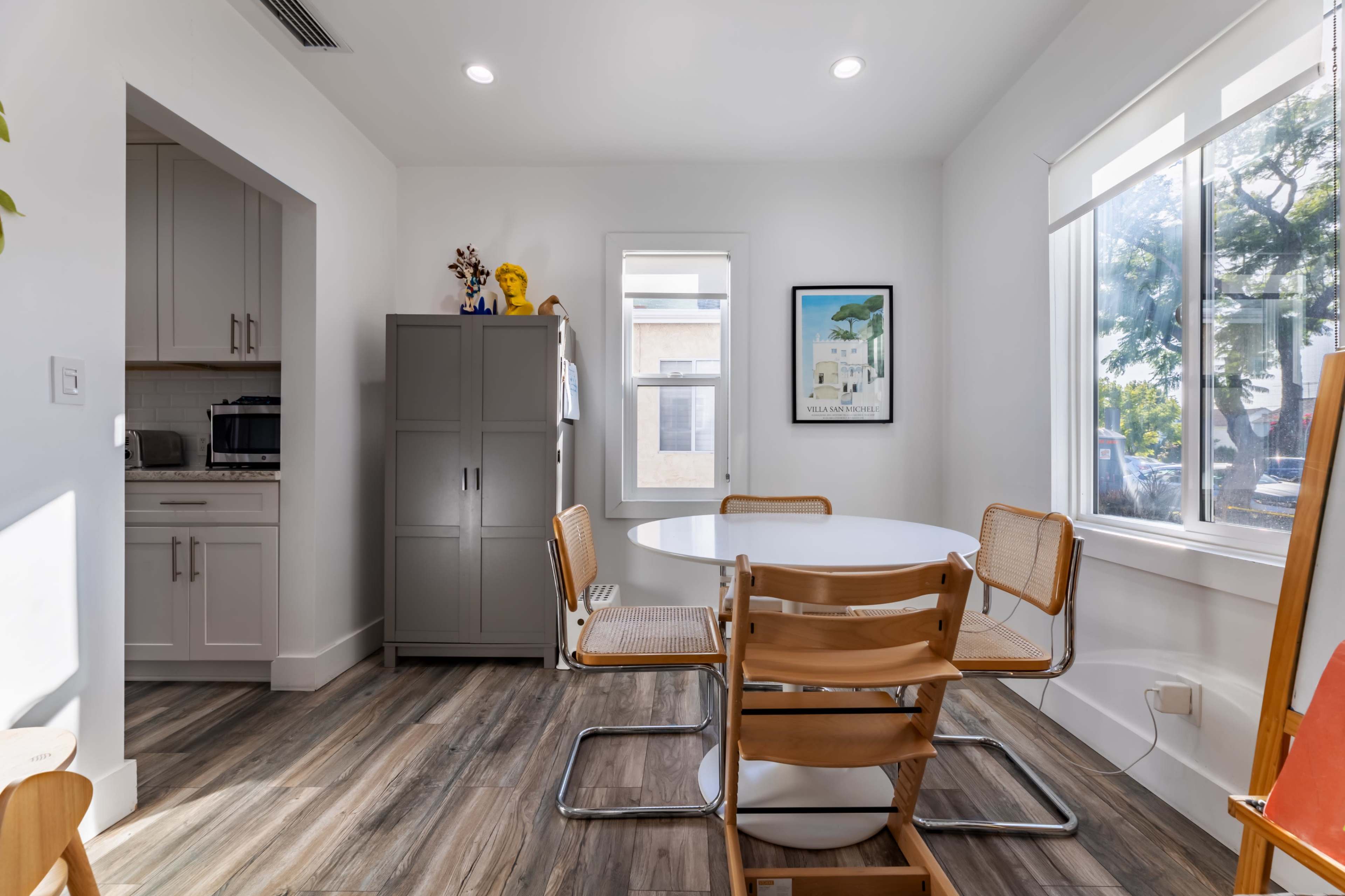 A modern dining area features a round white table surrounded by four wooden chairs, with a cabinet and kitchen visible in the background.