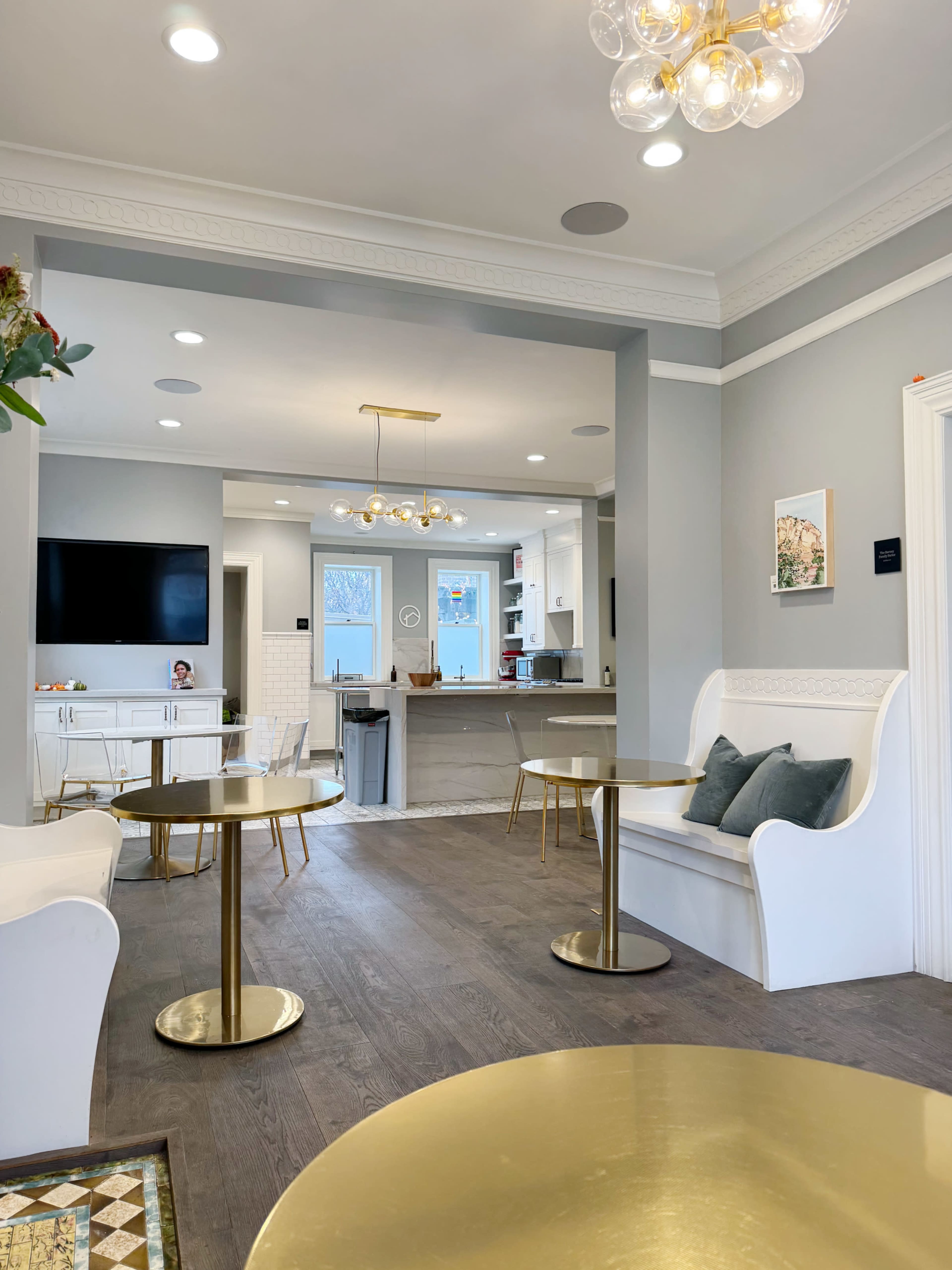 A modern, well-lit lounge area with white seating and brass tables, leading into a kitchen space.