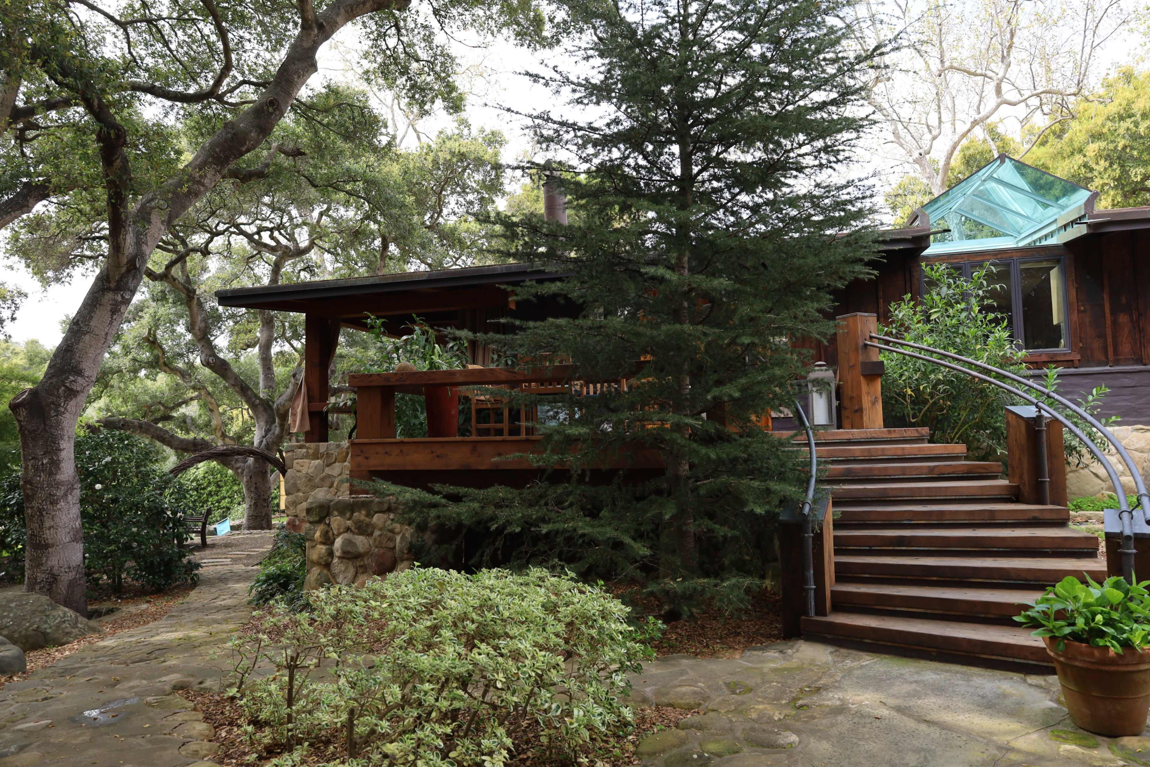 A wooden house with a stone foundation is surrounded by trees and features a set of wooden stairs leading to a porch.