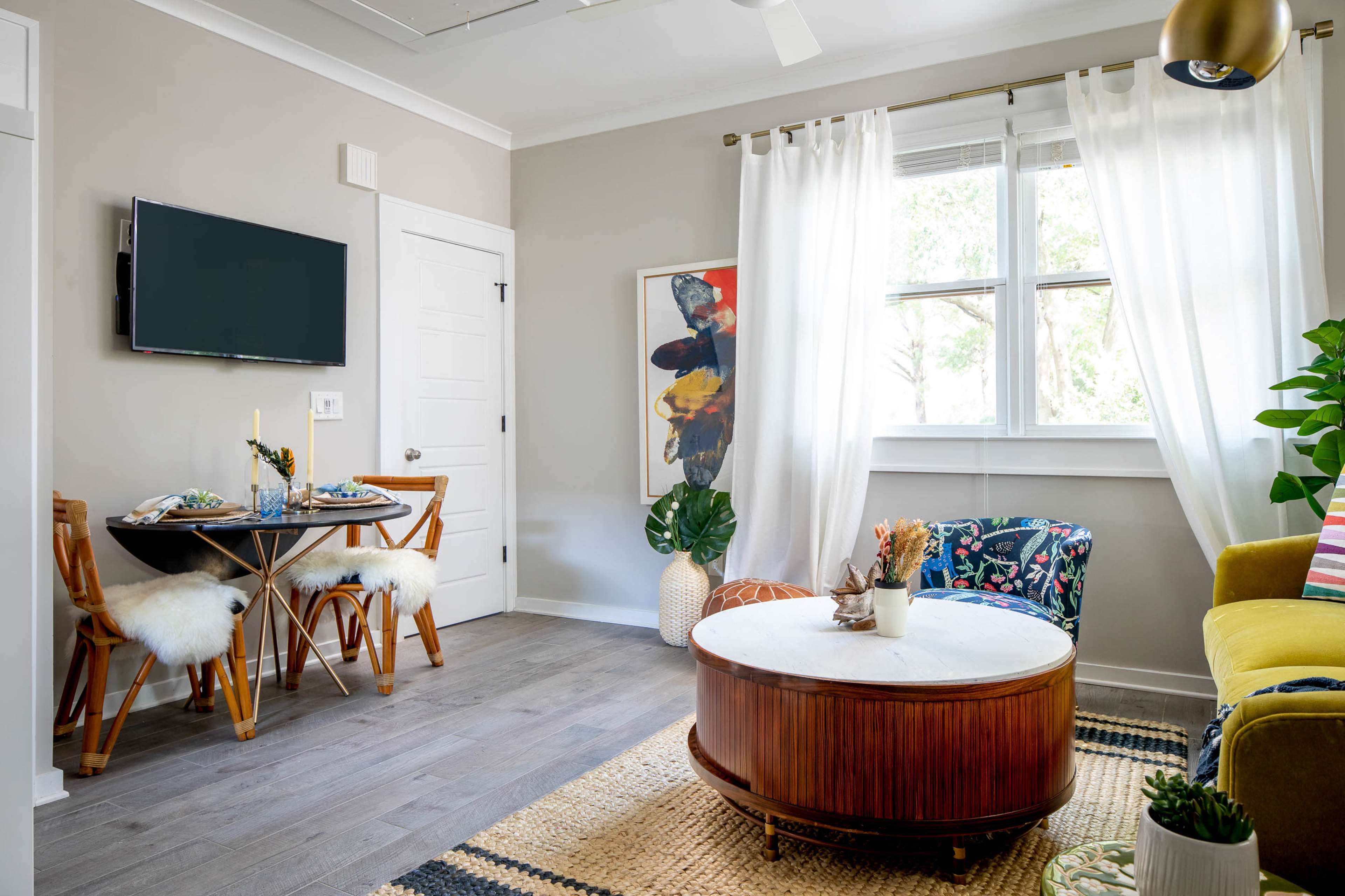 The image shows a cozy living space with a dining area featuring a round table and chairs, a colorful sofa, a coffee table, and large windows dressed with white curtains.