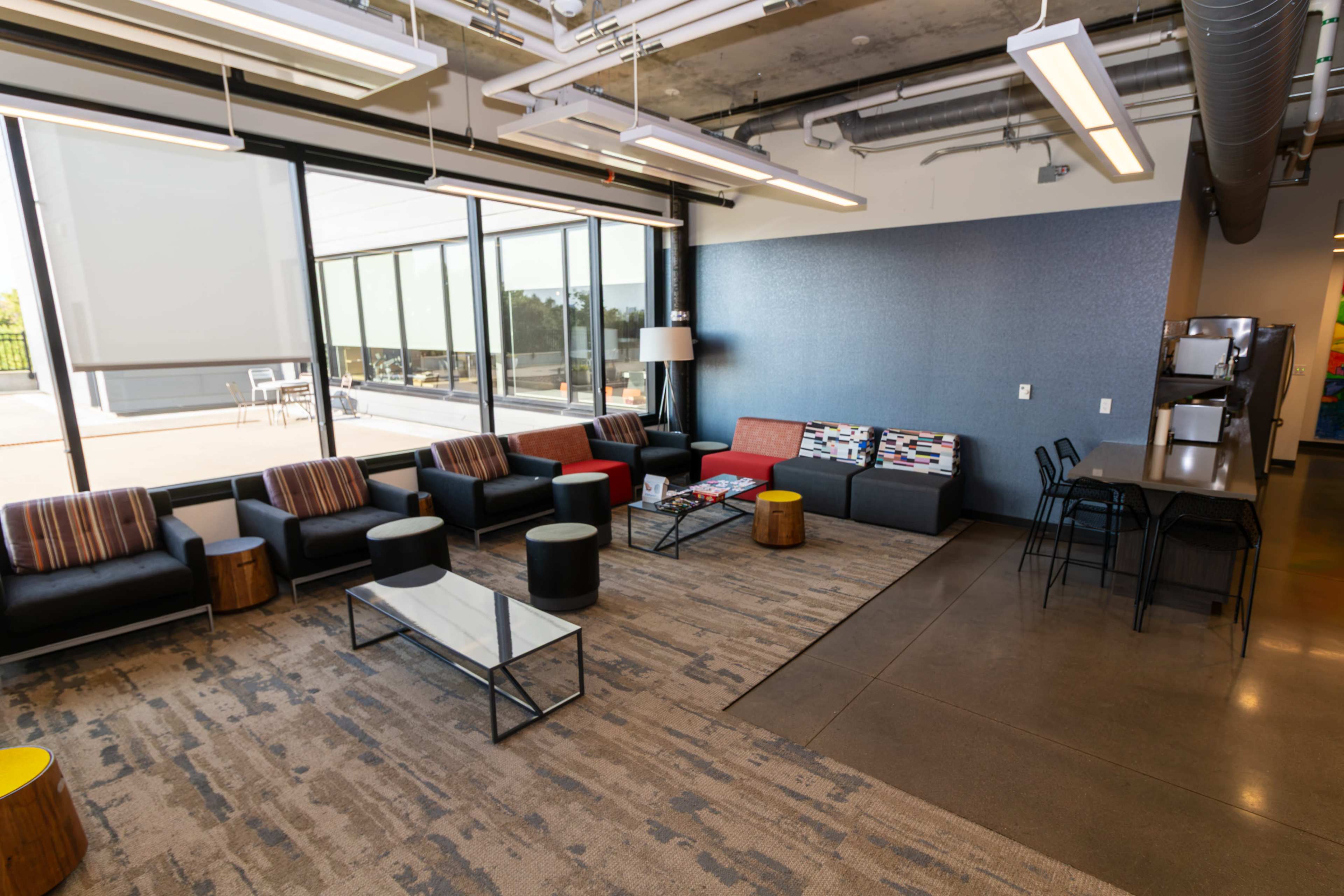 The image shows a modern lounge area with black sofas, wooden stools, and large windows allowing natural light to fill the space.