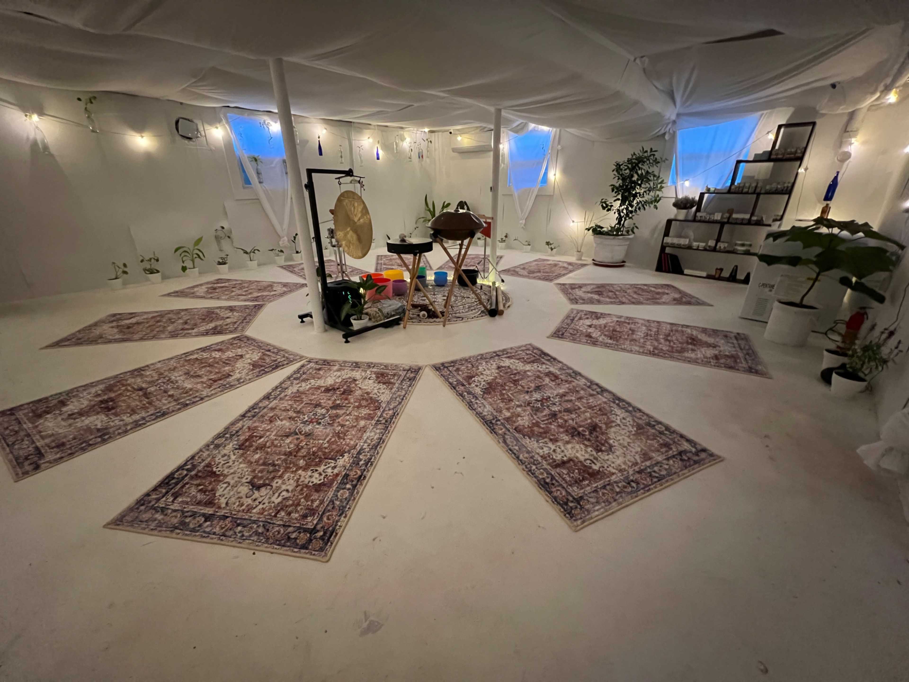 A spacious, softly lit room with an arrangement of rugs on the floor, a gong, a table with various items, and plants along the walls.