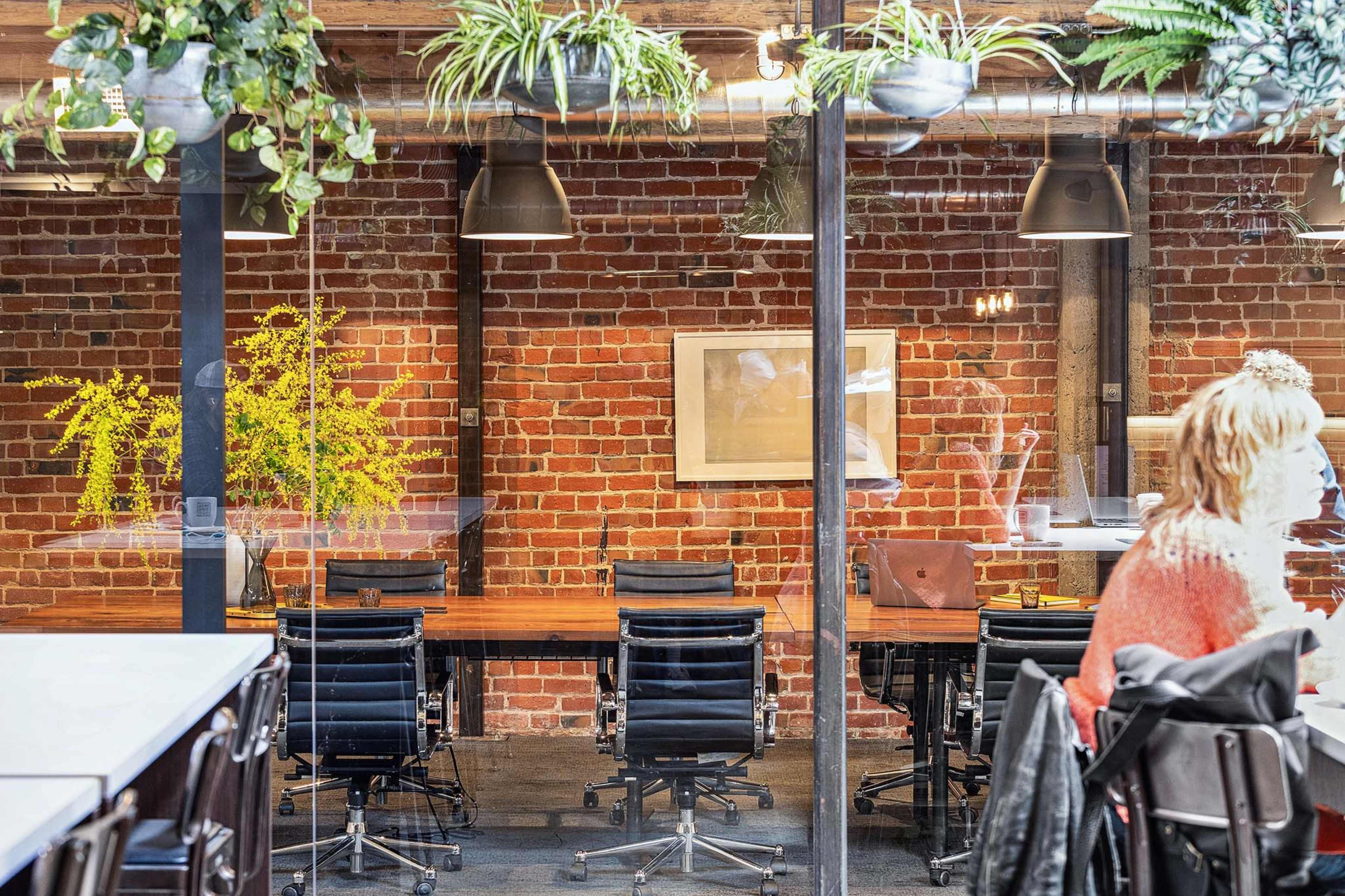 A modern office space with a brick wall, wooden tables, black chairs, and indoor plants.