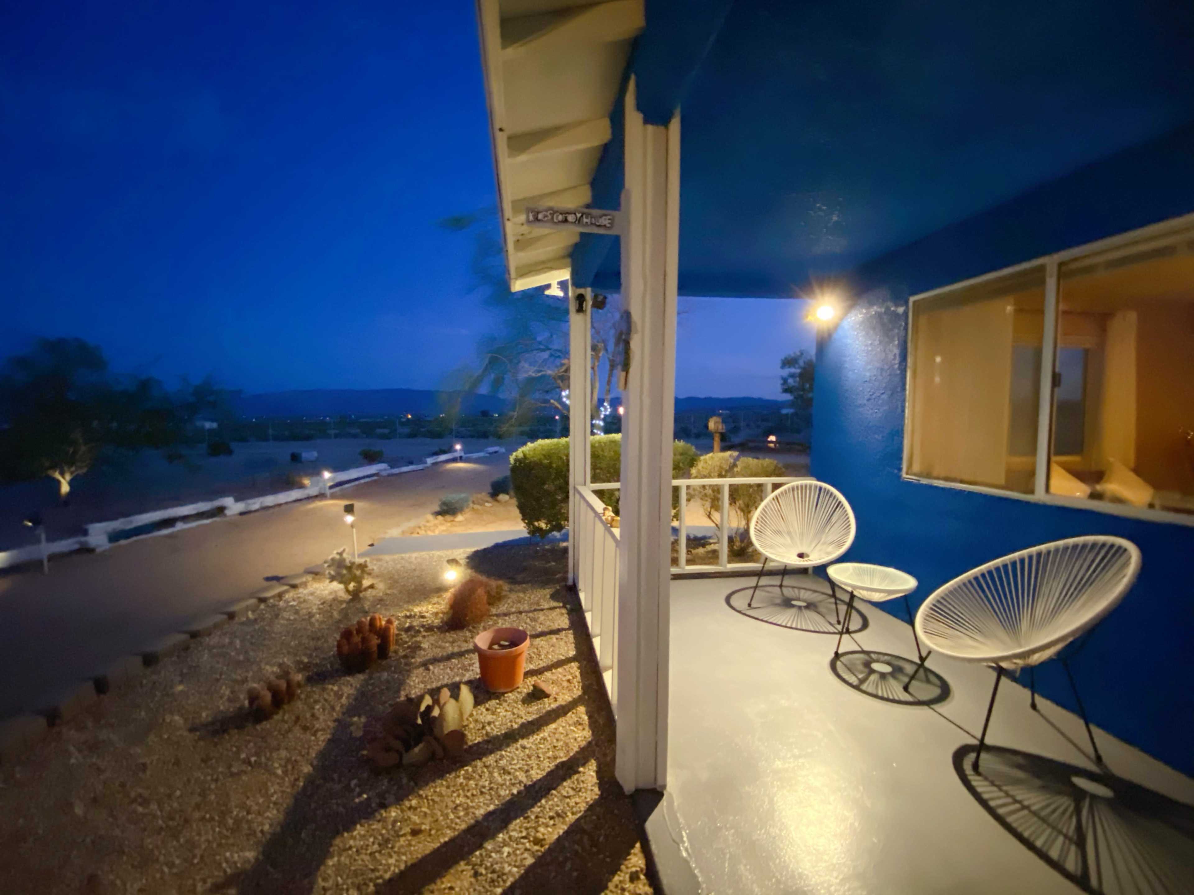 The image shows a cozy porch area with two chairs, illuminated by soft lighting, overlooking a gravel path at twilight.