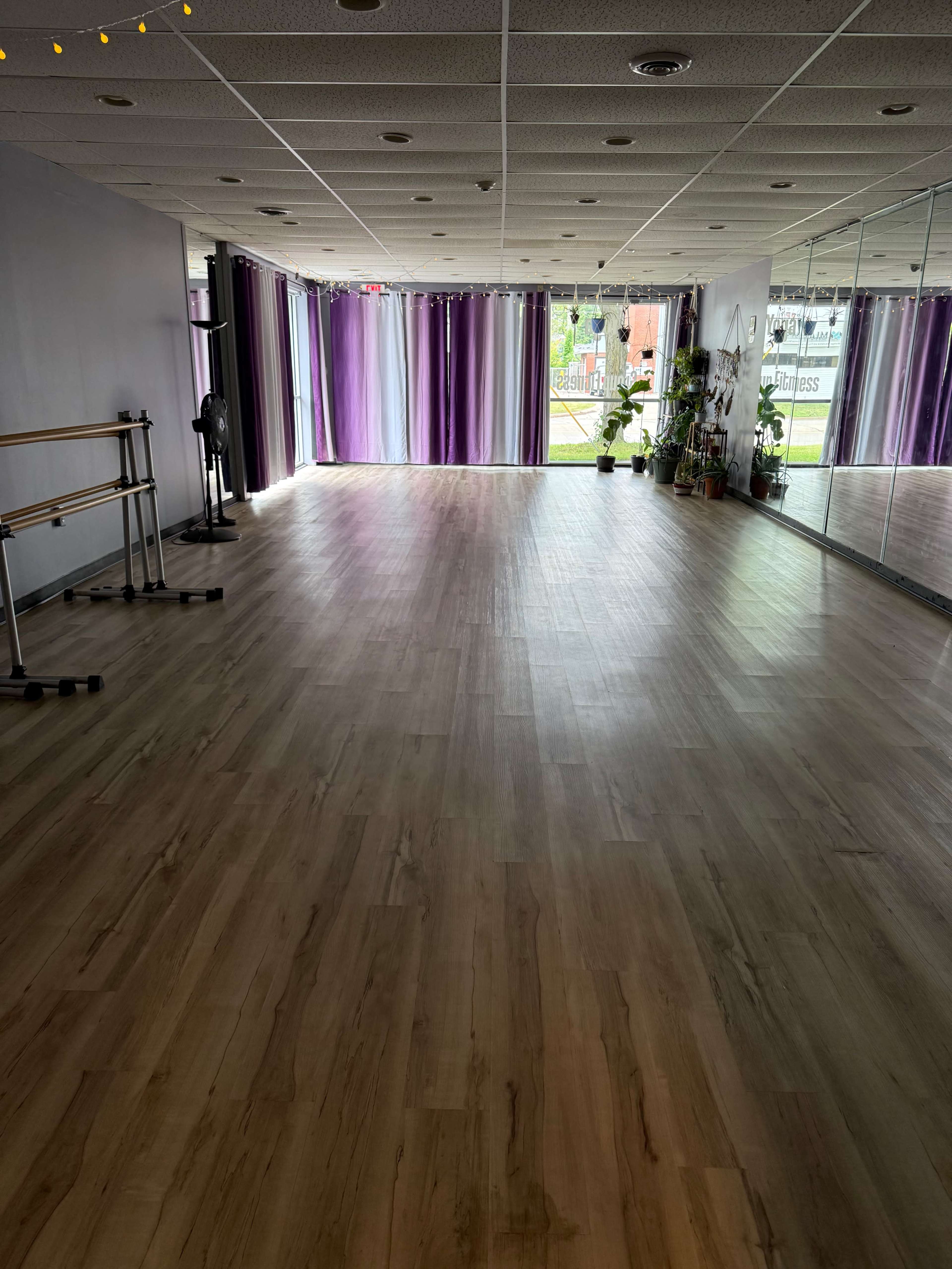 The image shows a spacious, well-lit dance studio with wooden flooring, large mirrors, and colorful curtains.