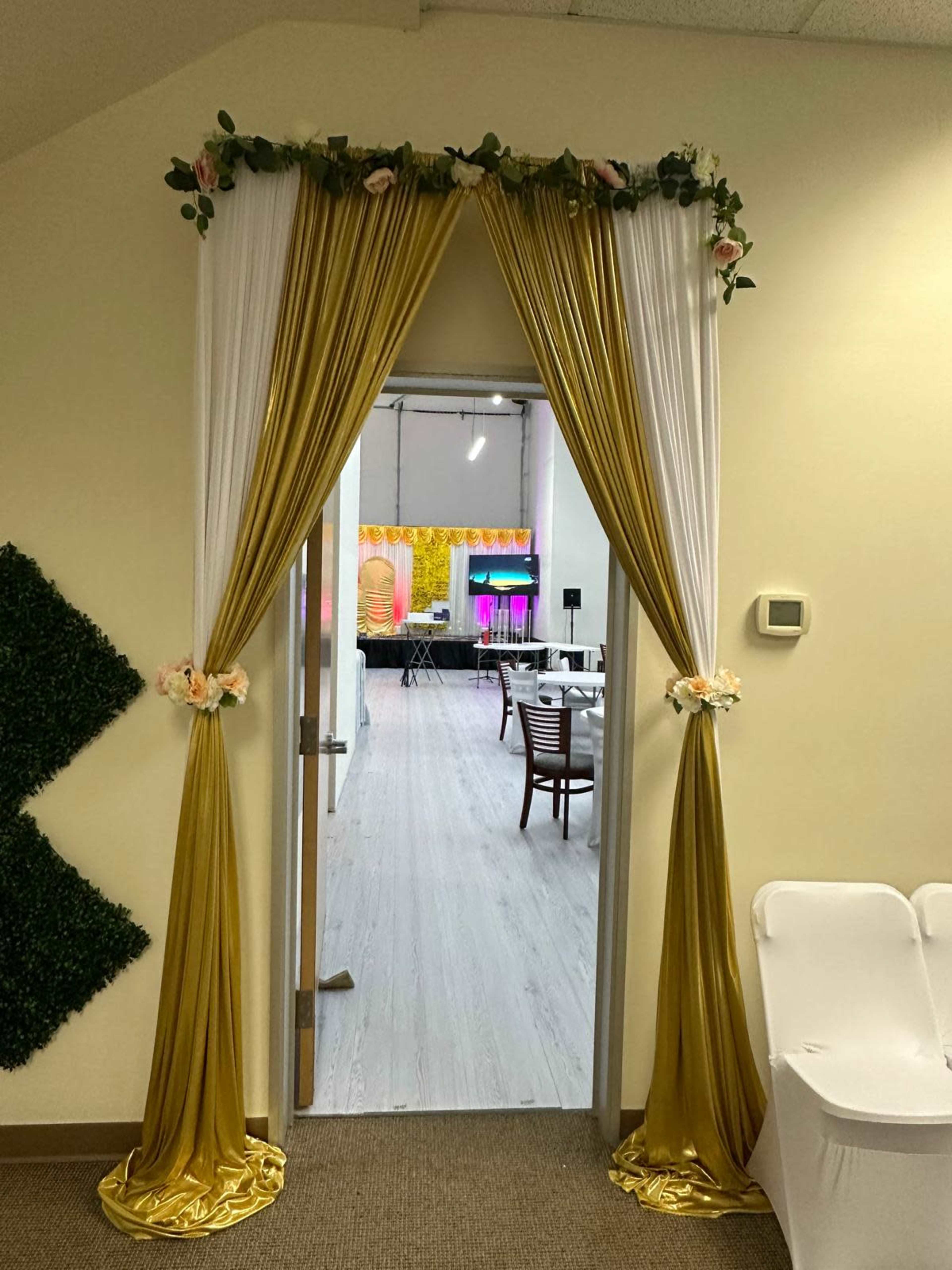 The image shows a doorway adorned with gold and white drapery, decorated with flowers and leaves, leading into a brightly lit event space.