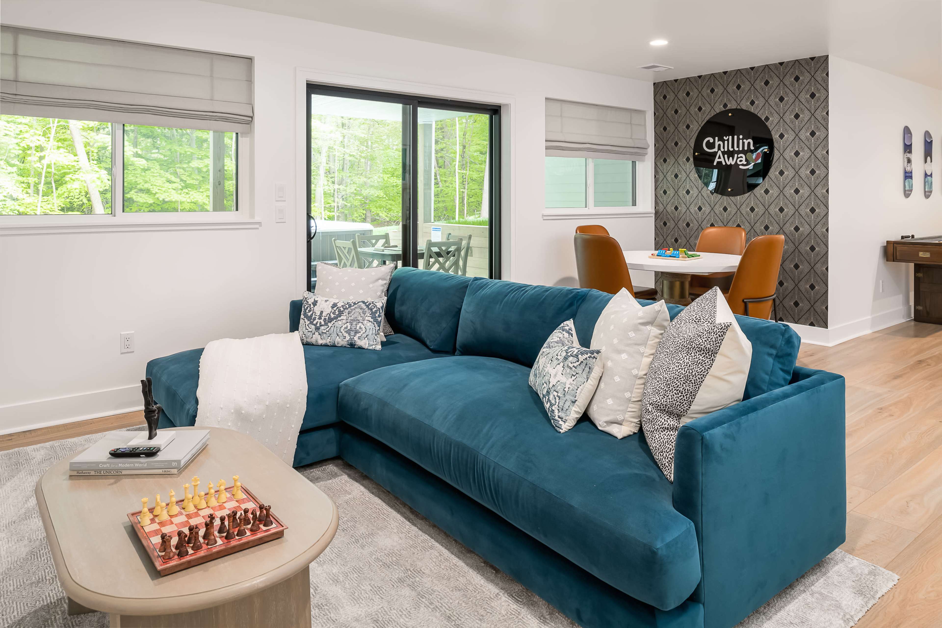 A modern living room features a teal sectional couch with decorative pillows, a coffee table with a chess set, and a dining area visible through large windows that overlook a forested landscape.