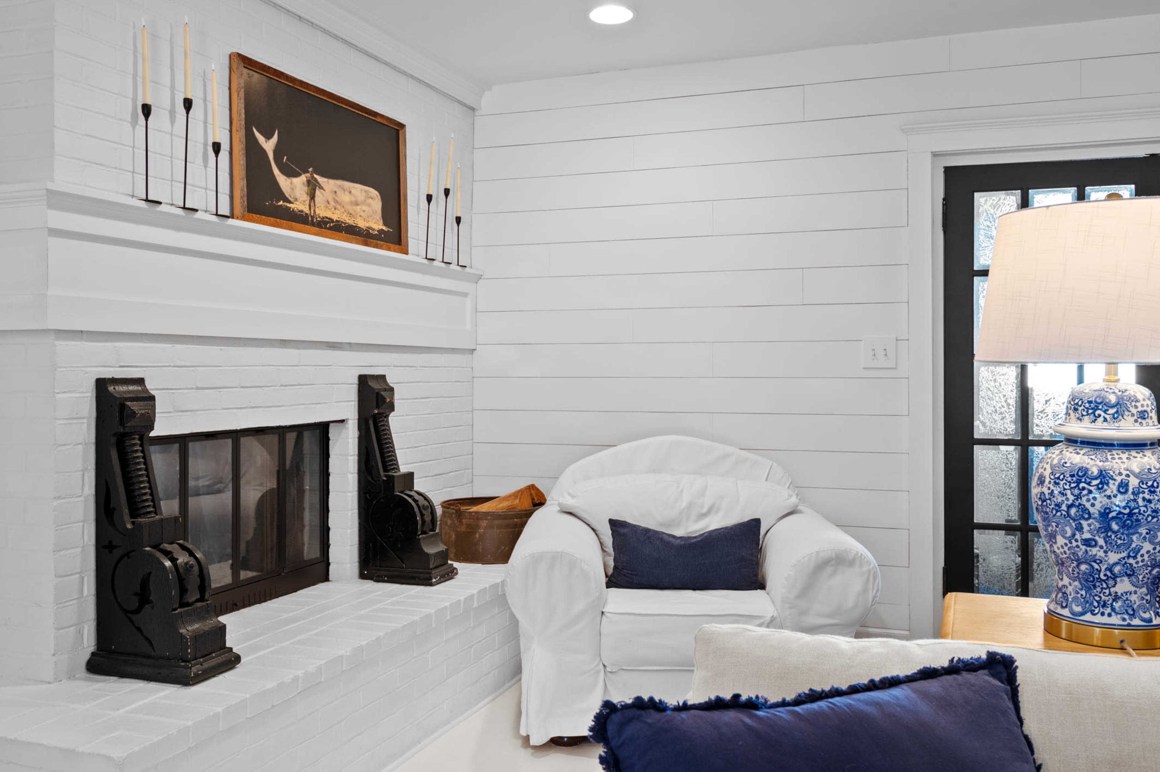 The image shows a cozy living room with a white fireplace, a comfortable white armchair, and a decorative lamp beside a blue and white vase.