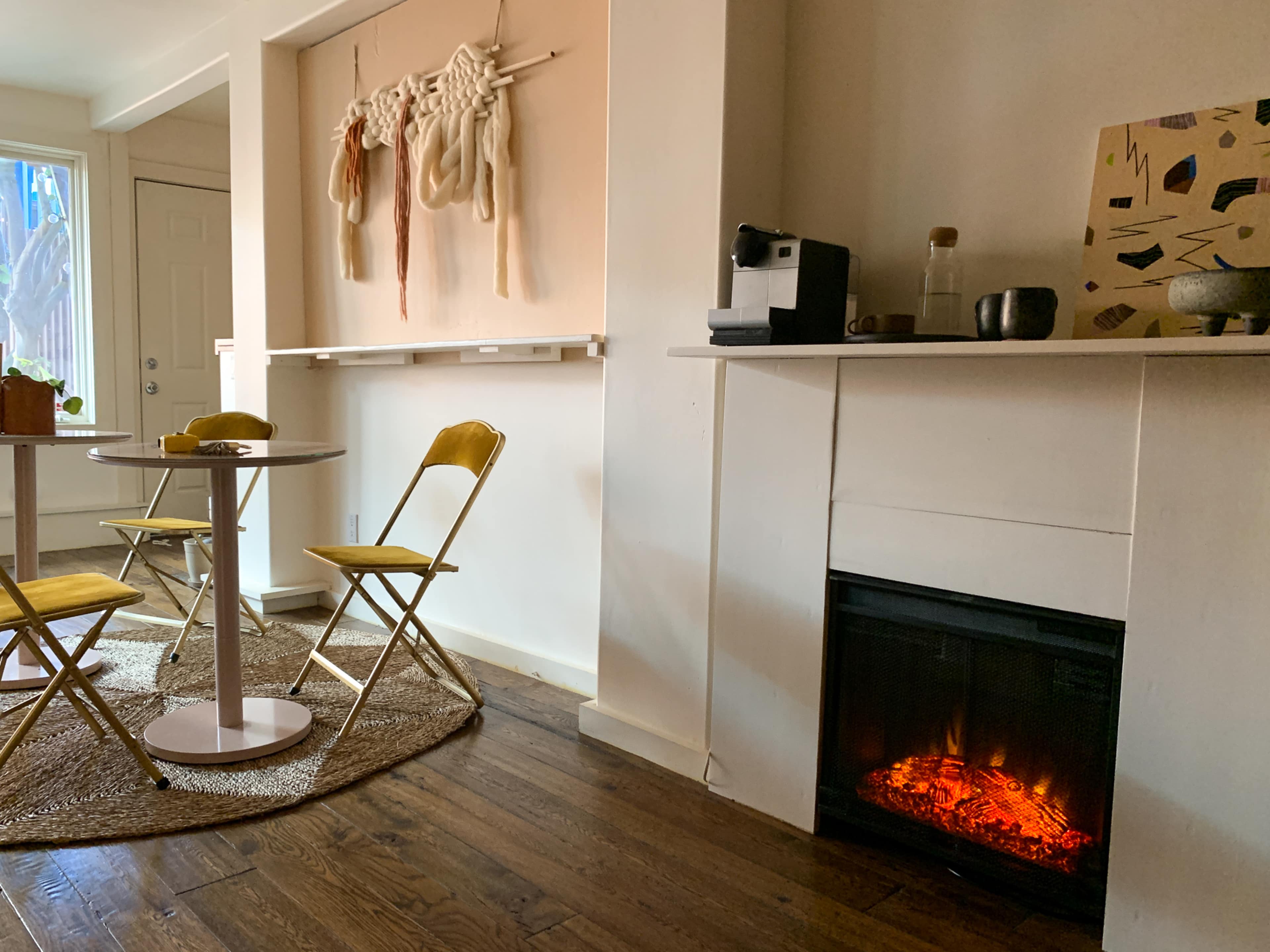 A cozy interior features a round table with yellow chairs, a faux fireplace, and a wall adorned with decorative textiles.