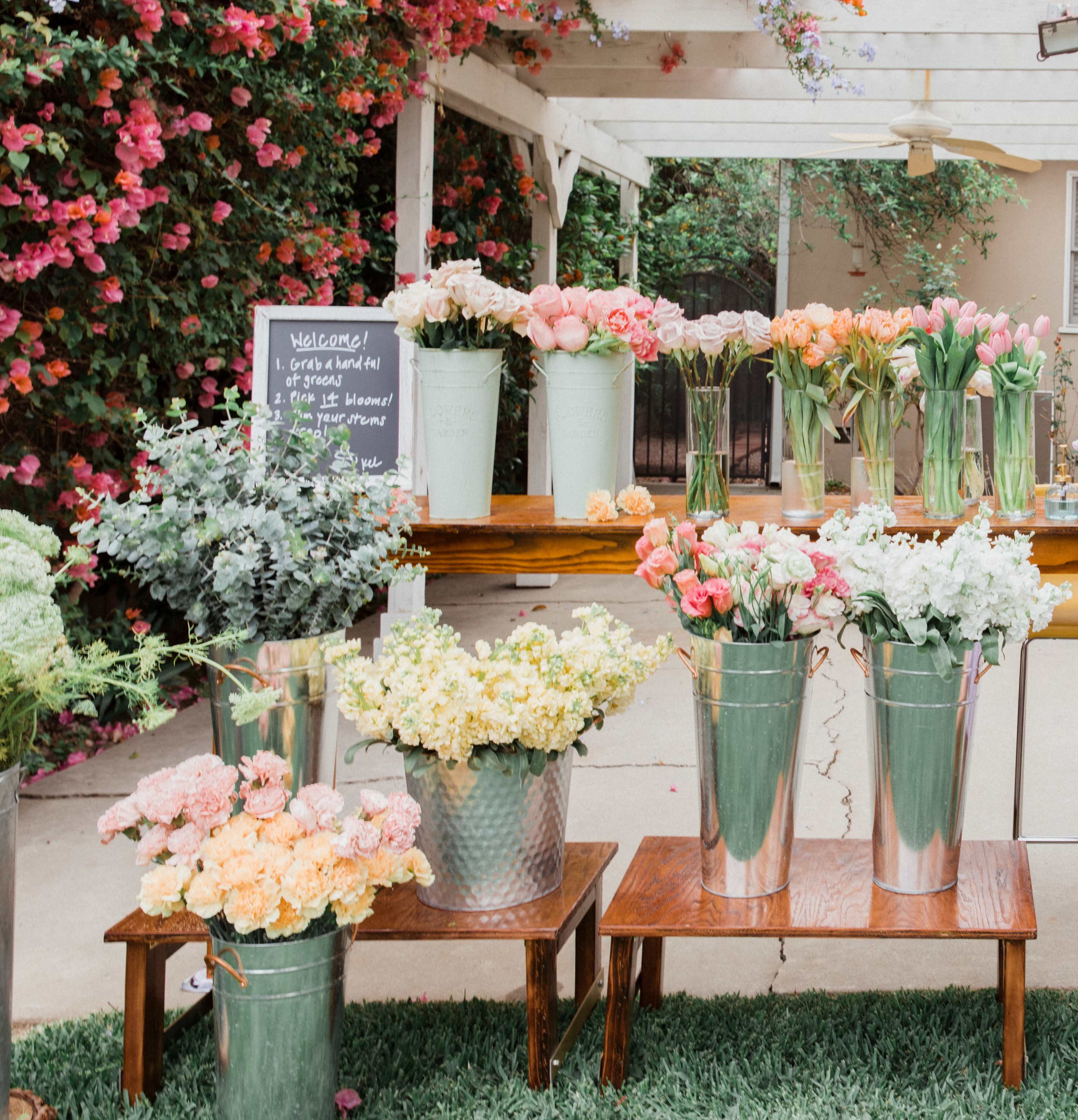 A display of various flowers in metal buckets is arranged on wooden tables under a white gazebo adorned with colorful blooms.