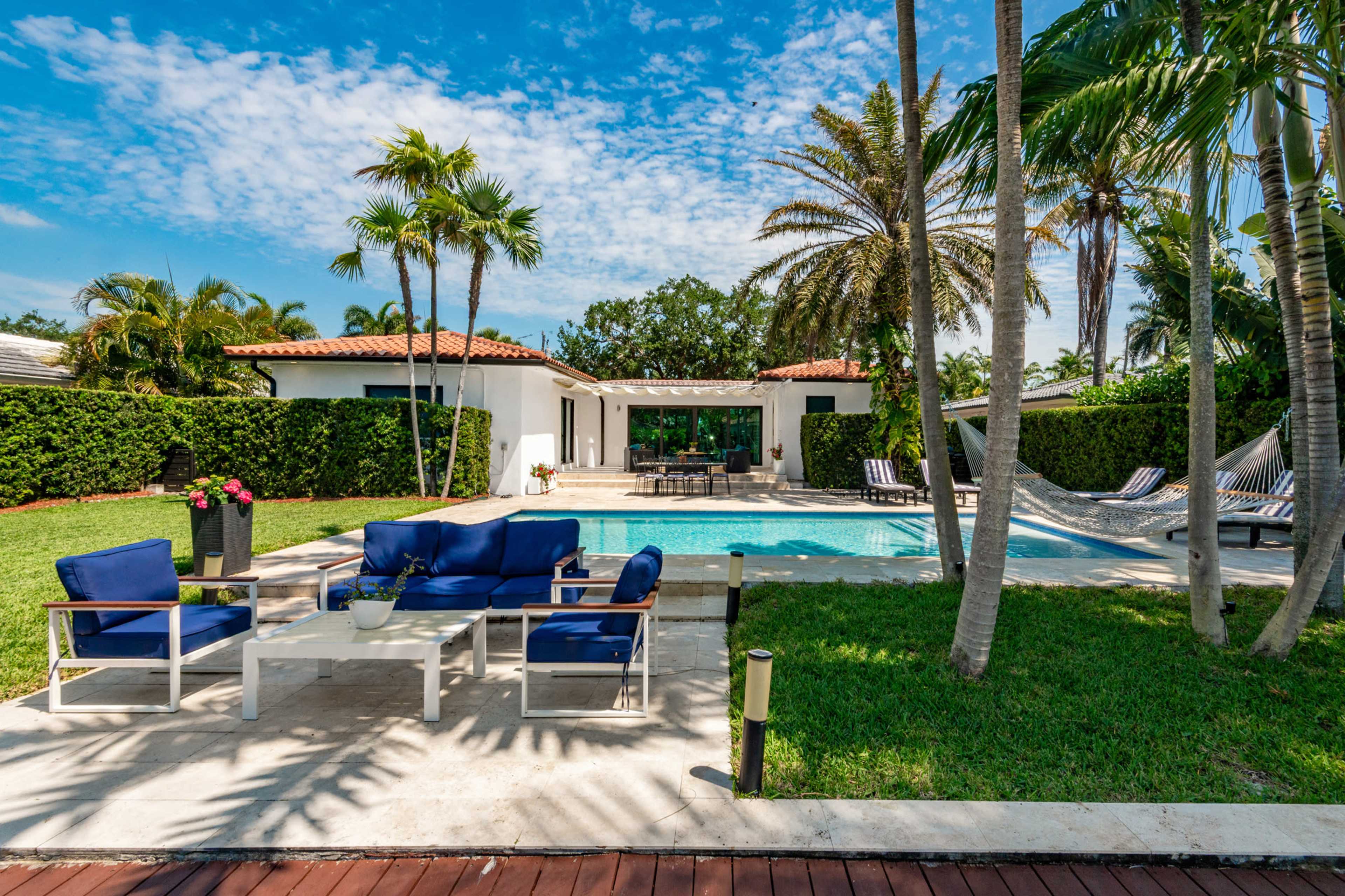 The image shows a poolside area at a residence, featuring lounge chairs, a table, and a swimming pool surrounded by palm trees and manicured grass.
