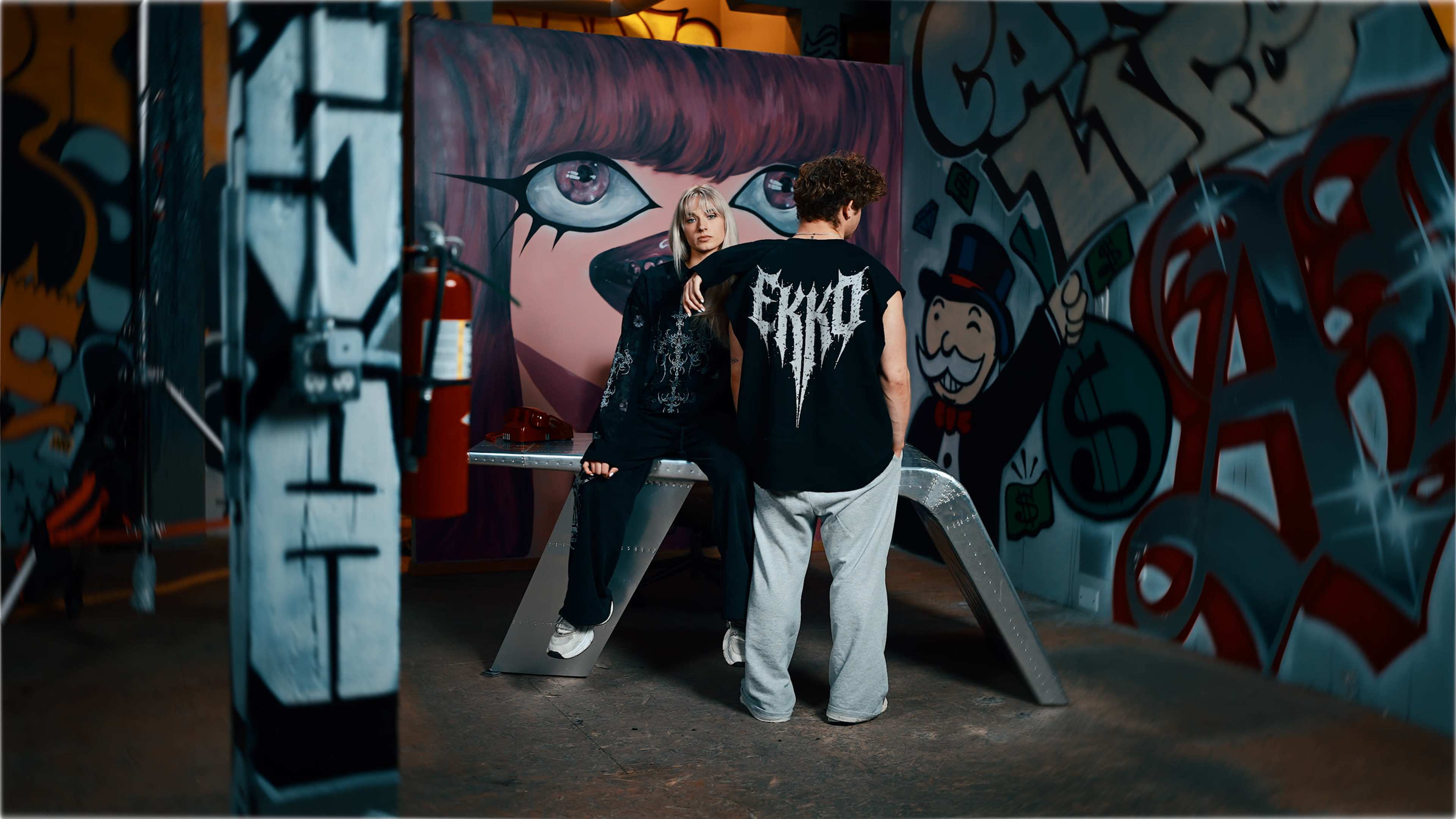 Two individuals stand in front of a colorful graffiti mural, one seated on a metallic bench while the other leans against it, both wearing streetwear.