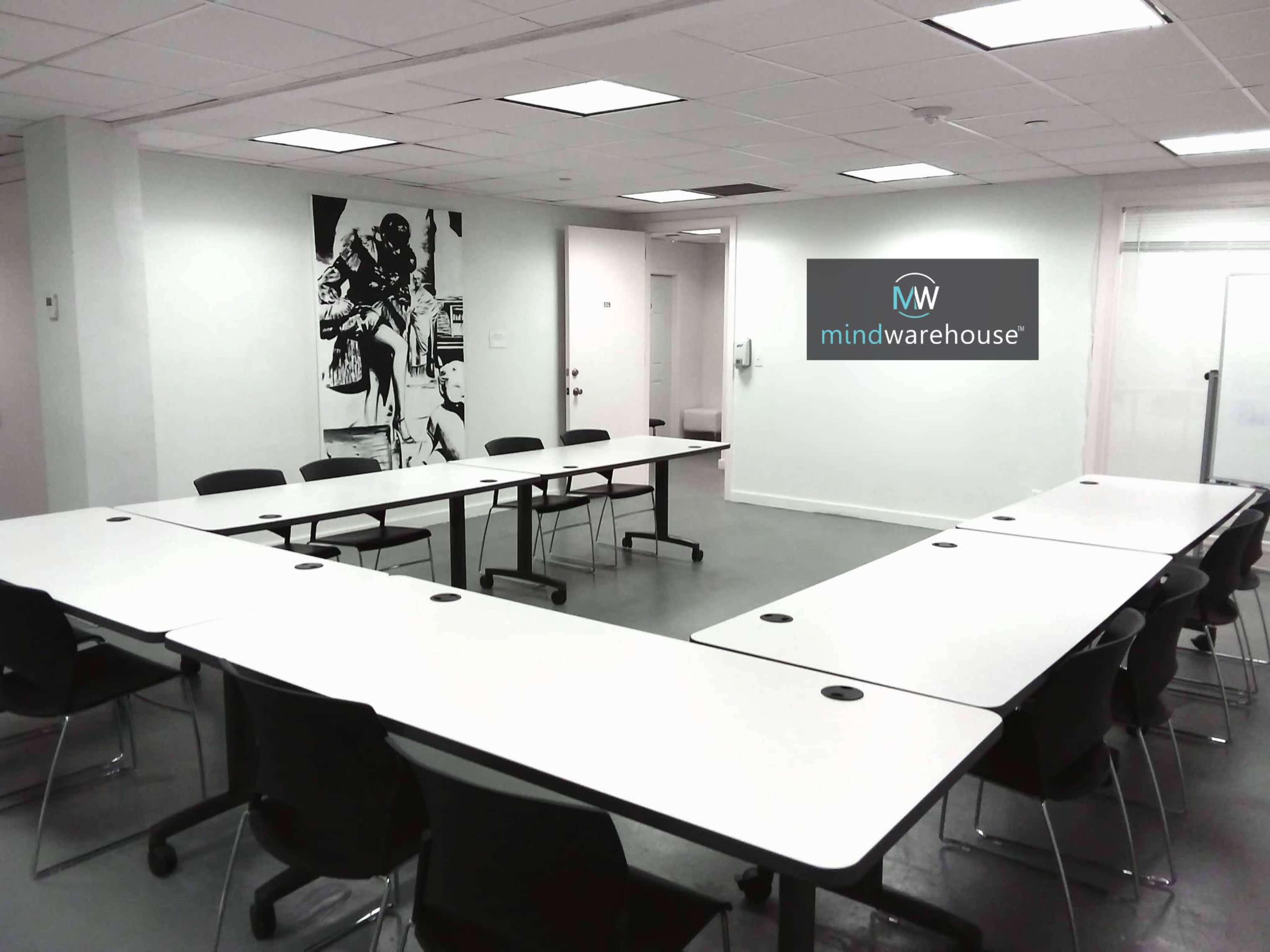A meeting room features a U-shaped arrangement of white tables and black chairs, with artwork on one wall and a logo on another.