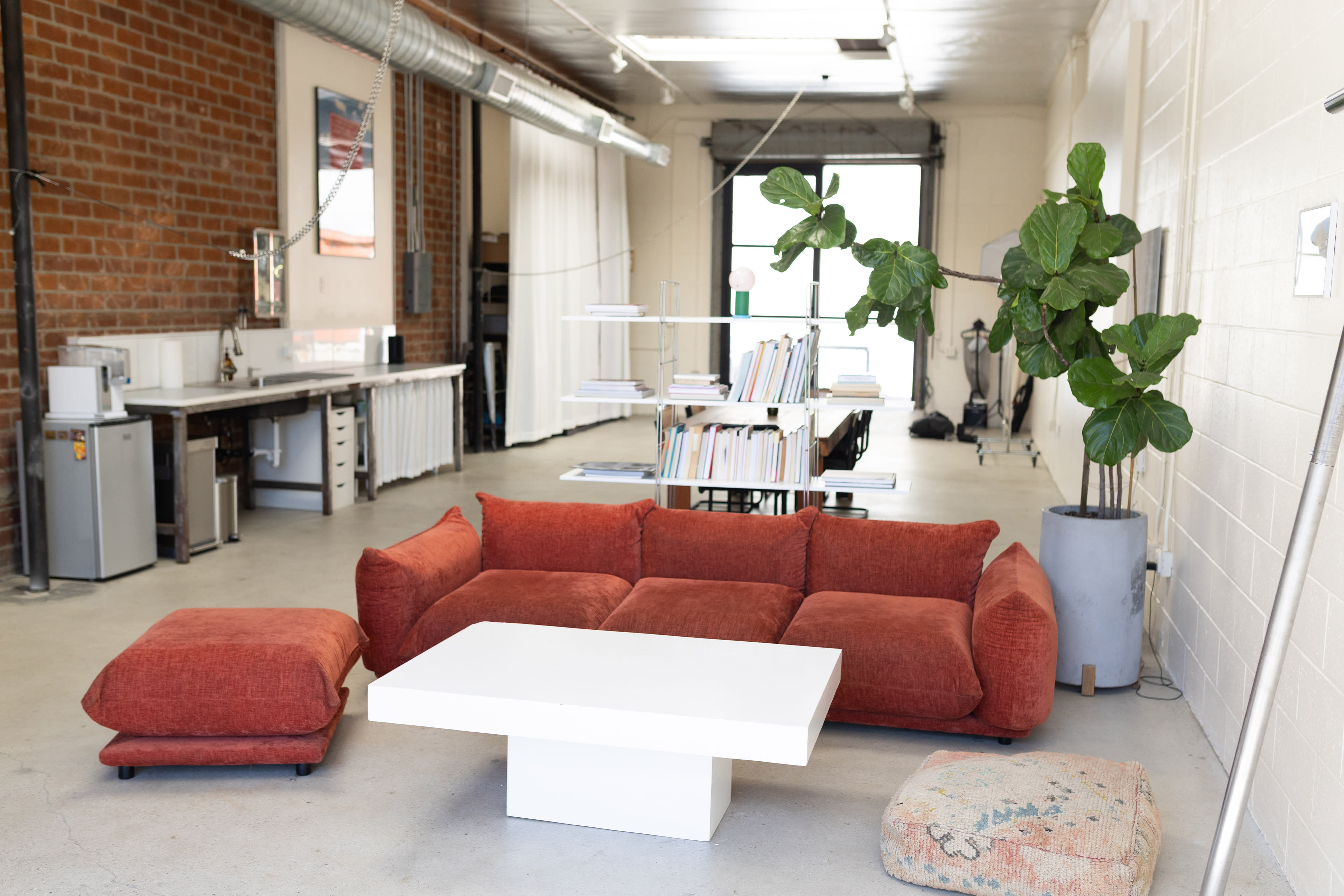 The image shows a modern, open workspace featuring a red sectional sofa, a white coffee table, and a large potted plant, with various office supplies and equipment visible in the background.