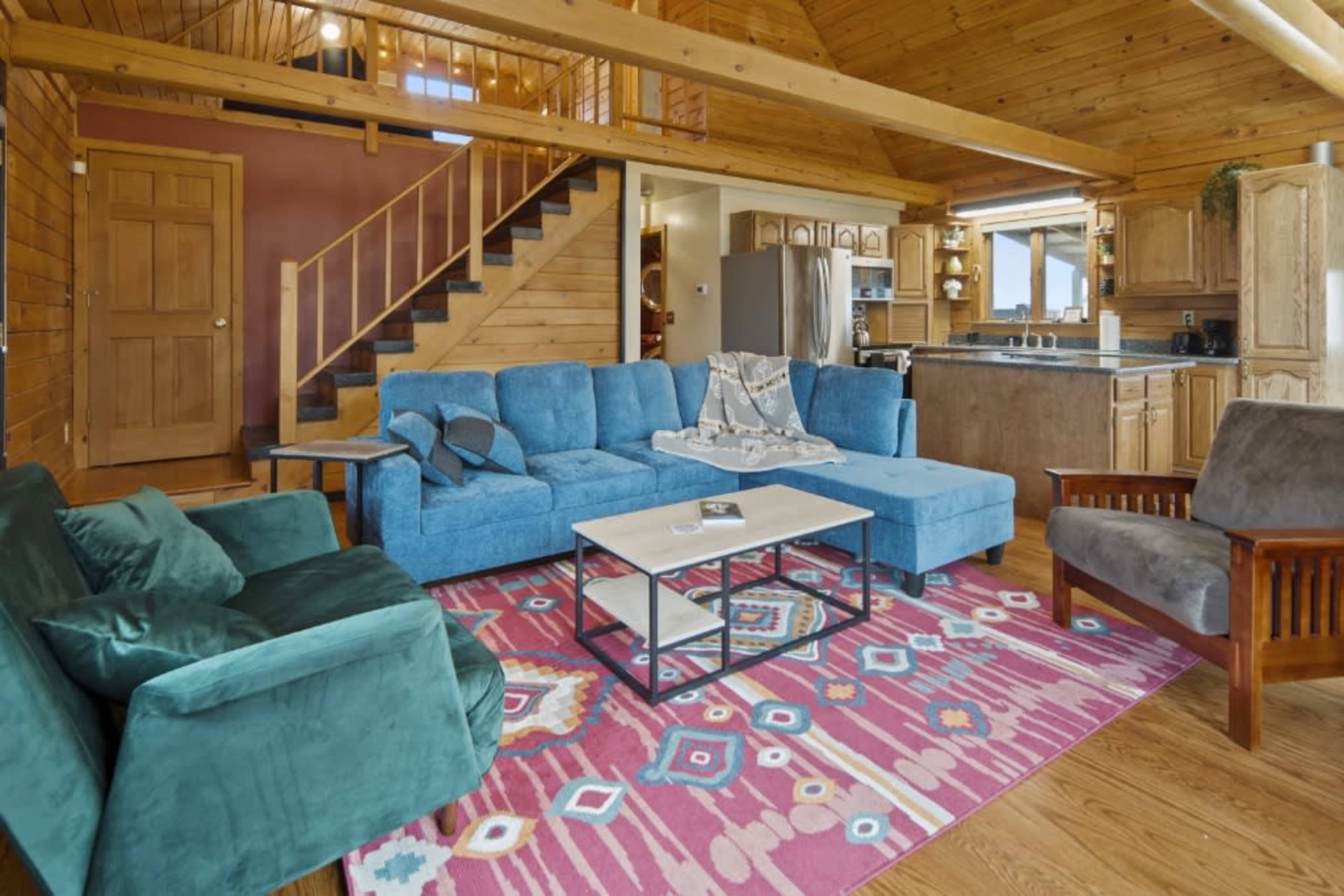 The room features a spacious living area with a sectional sofa, a coffee table, and a colorful area rug, all set against a backdrop of wooden walls and a staircase leading to an upper level.