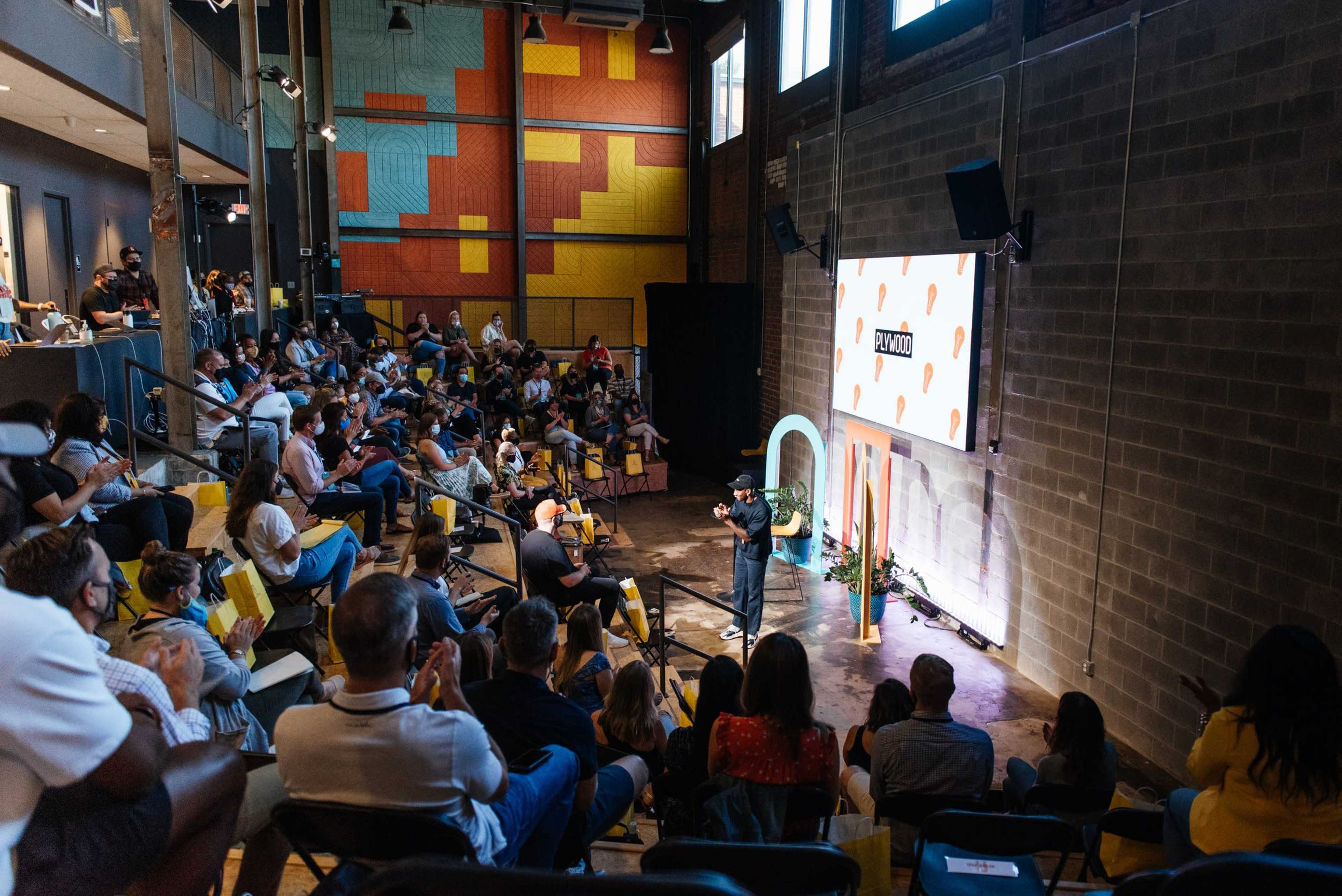 A speaker stands on a stage in front of a large audience seated in a spacious venue with colorful wall art.