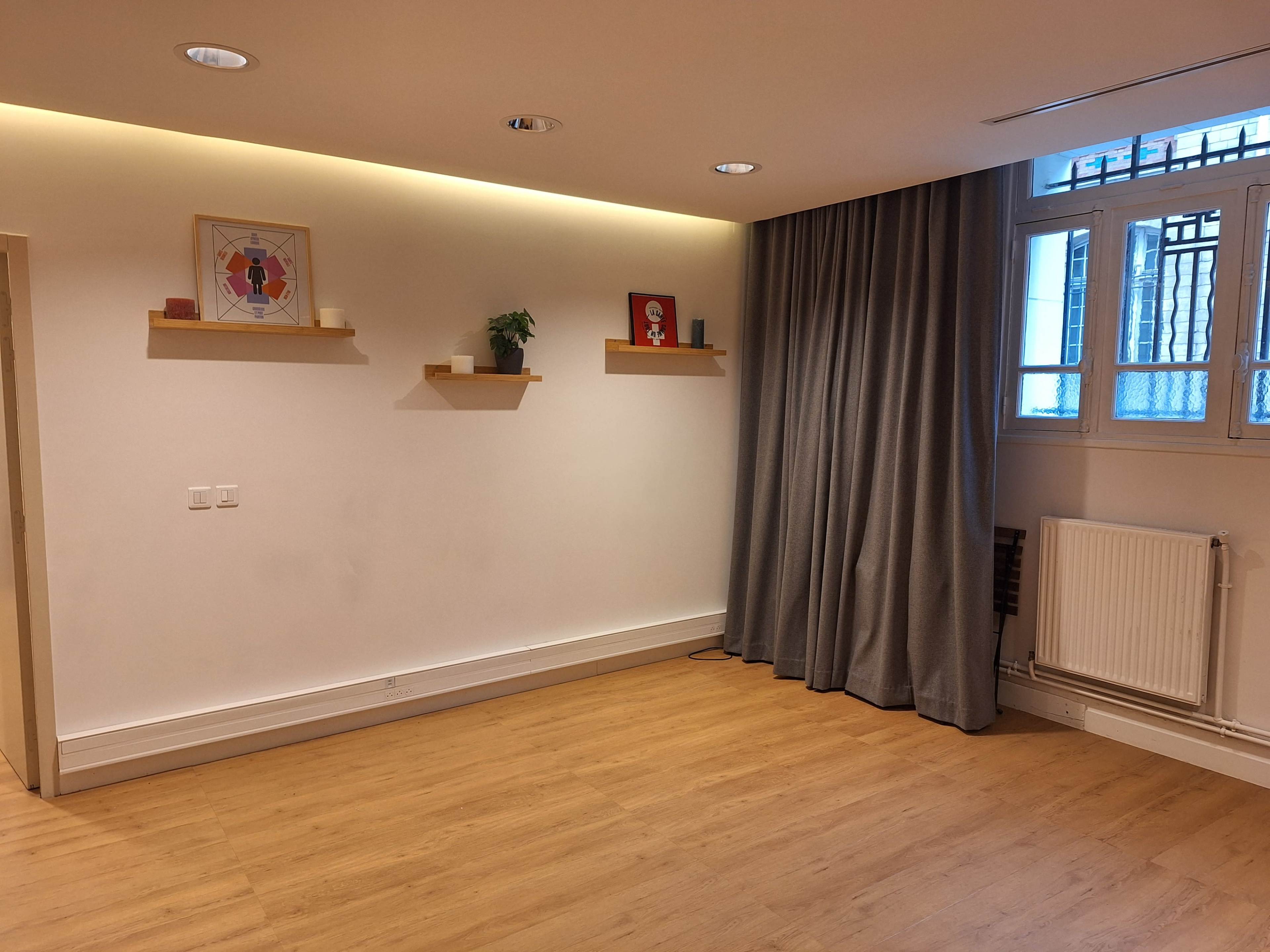 The room features light wooden flooring, white walls, two wall-mounted shelves with small plants and decorations, a gray curtain, and a radiator beneath a window with multiple panes.