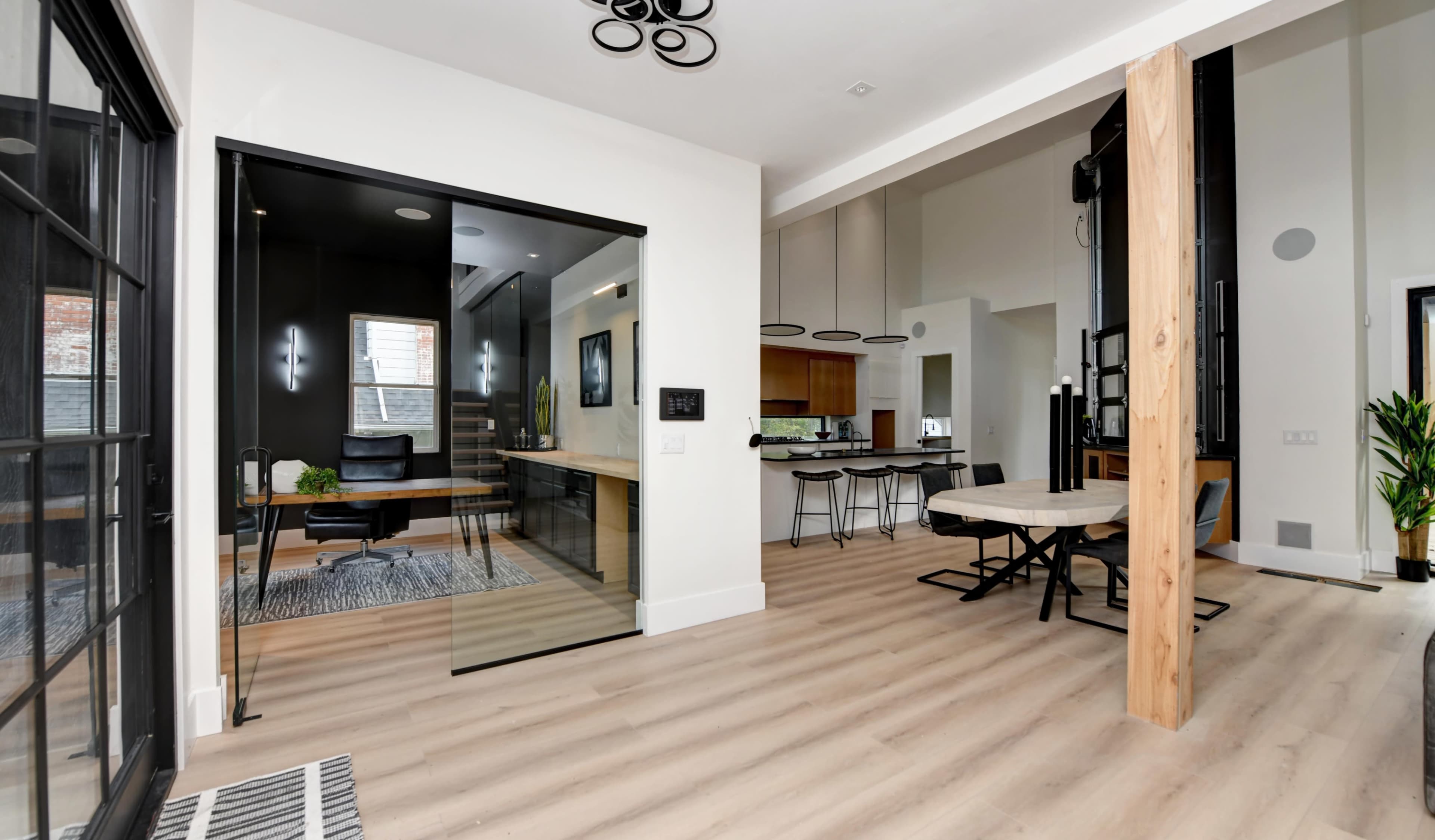 A modern interior space with an open layout featuring a dining area, a living area with wooden floors, and a separate workspace visible through a glass wall.