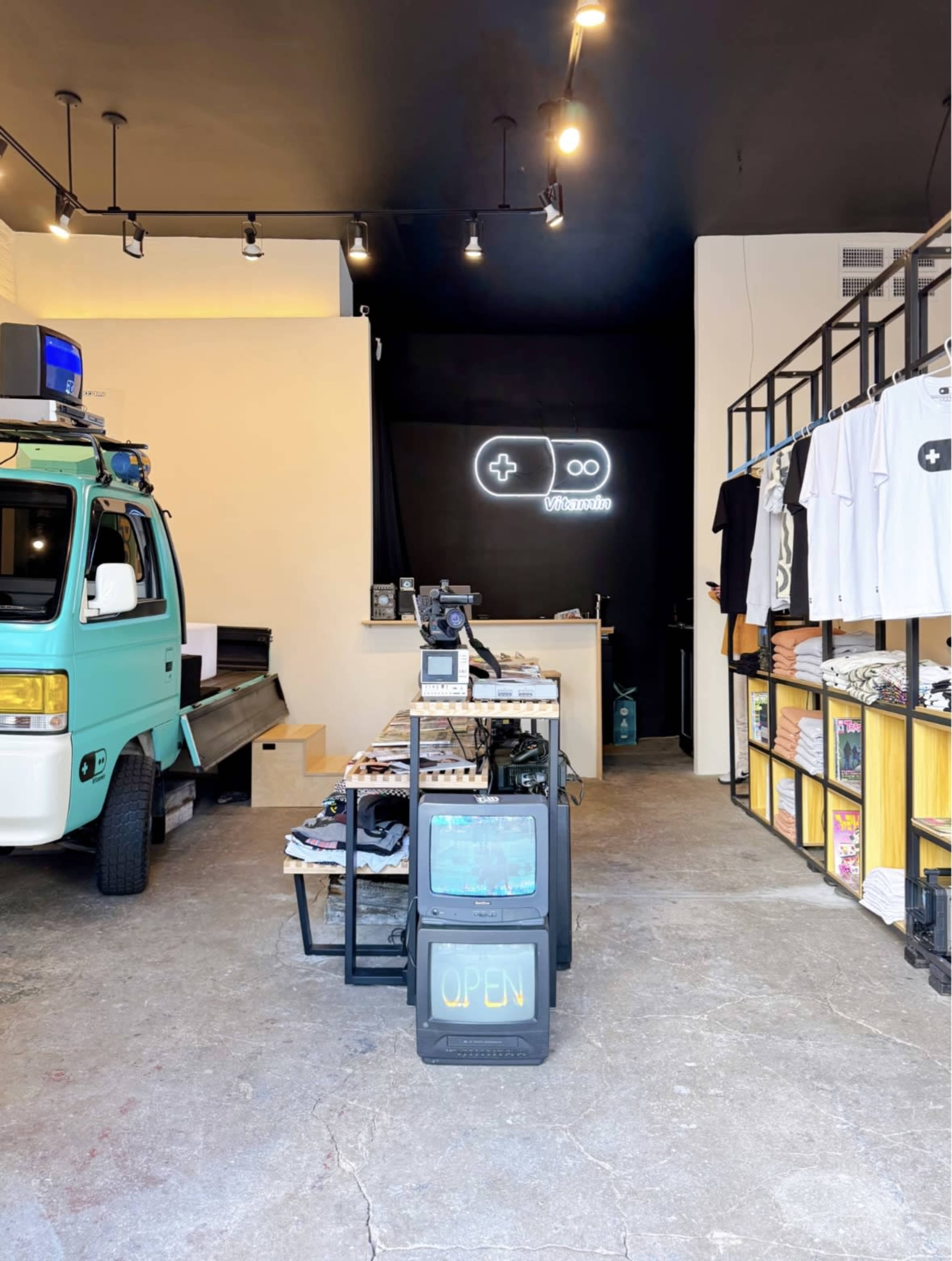 The interior of a retail store features a mint-green van, a display table with various merchandise, and a neon sign on the wall that resembles a game controller.