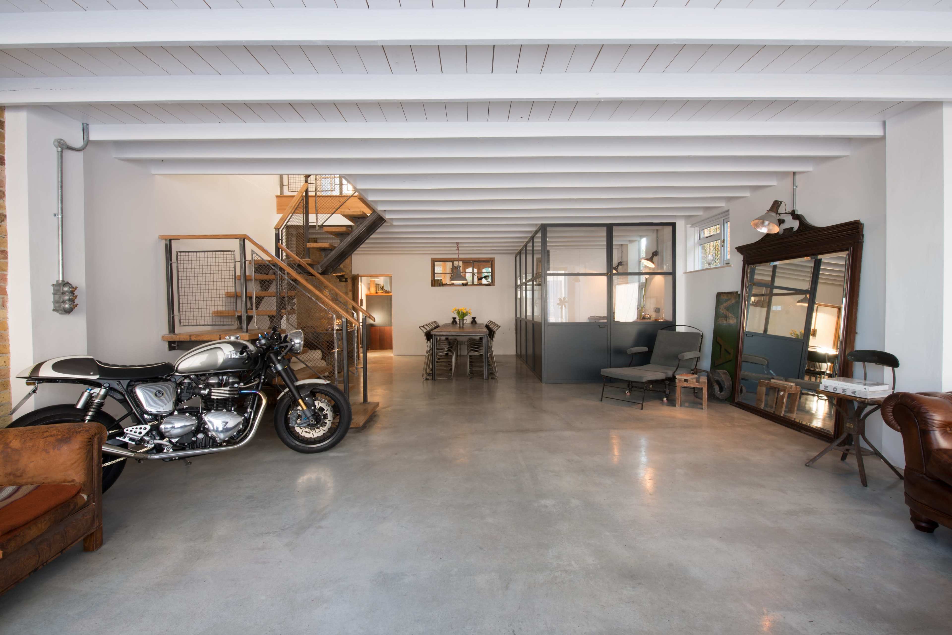 The image shows a spacious, industrial-style interior featuring a motorcycle, a wooden staircase, a dining area, and large windows.