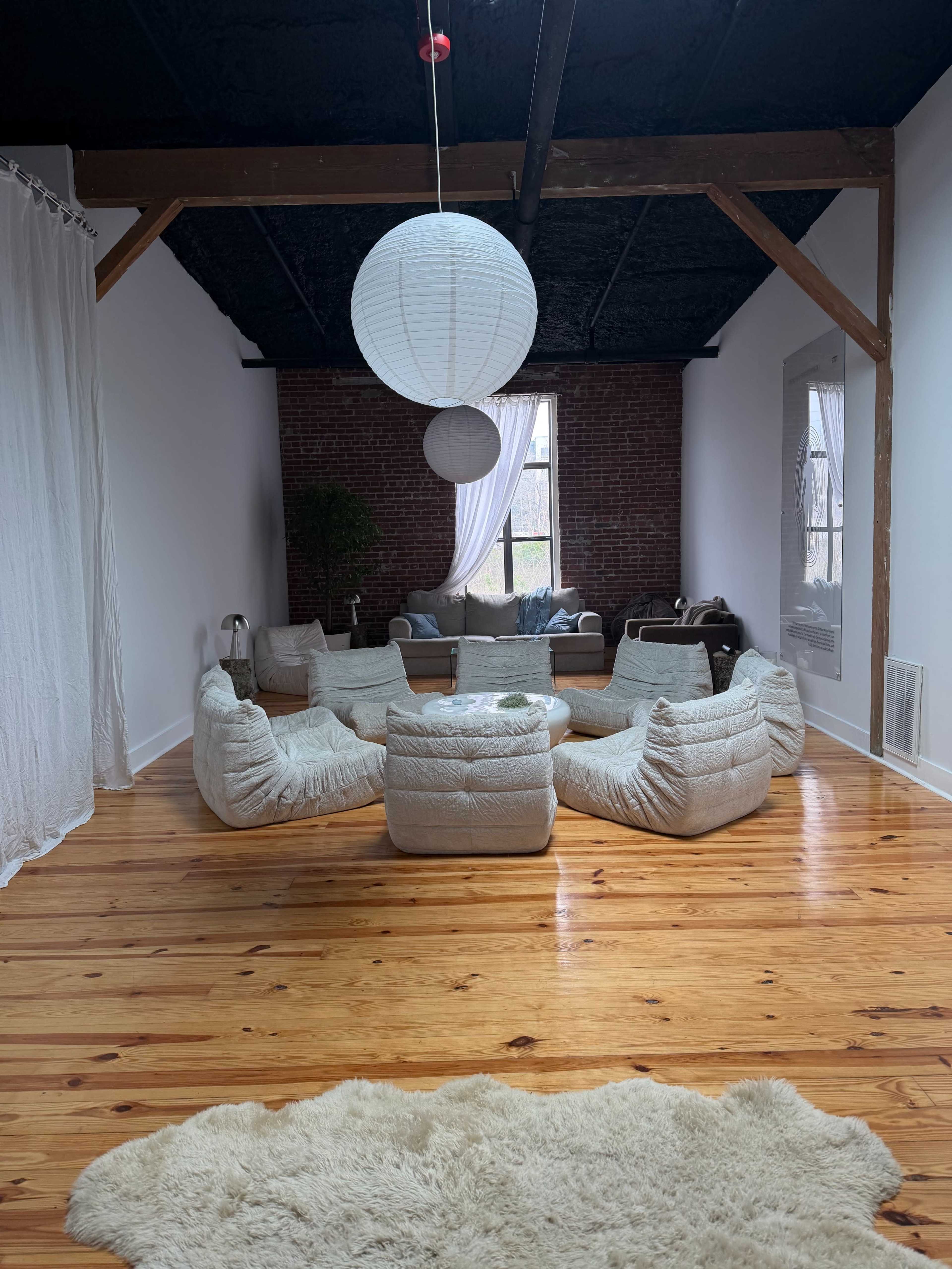 A spacious living area features a circular arrangement of cushioned seating, wooden floors, and large windows with natural light.