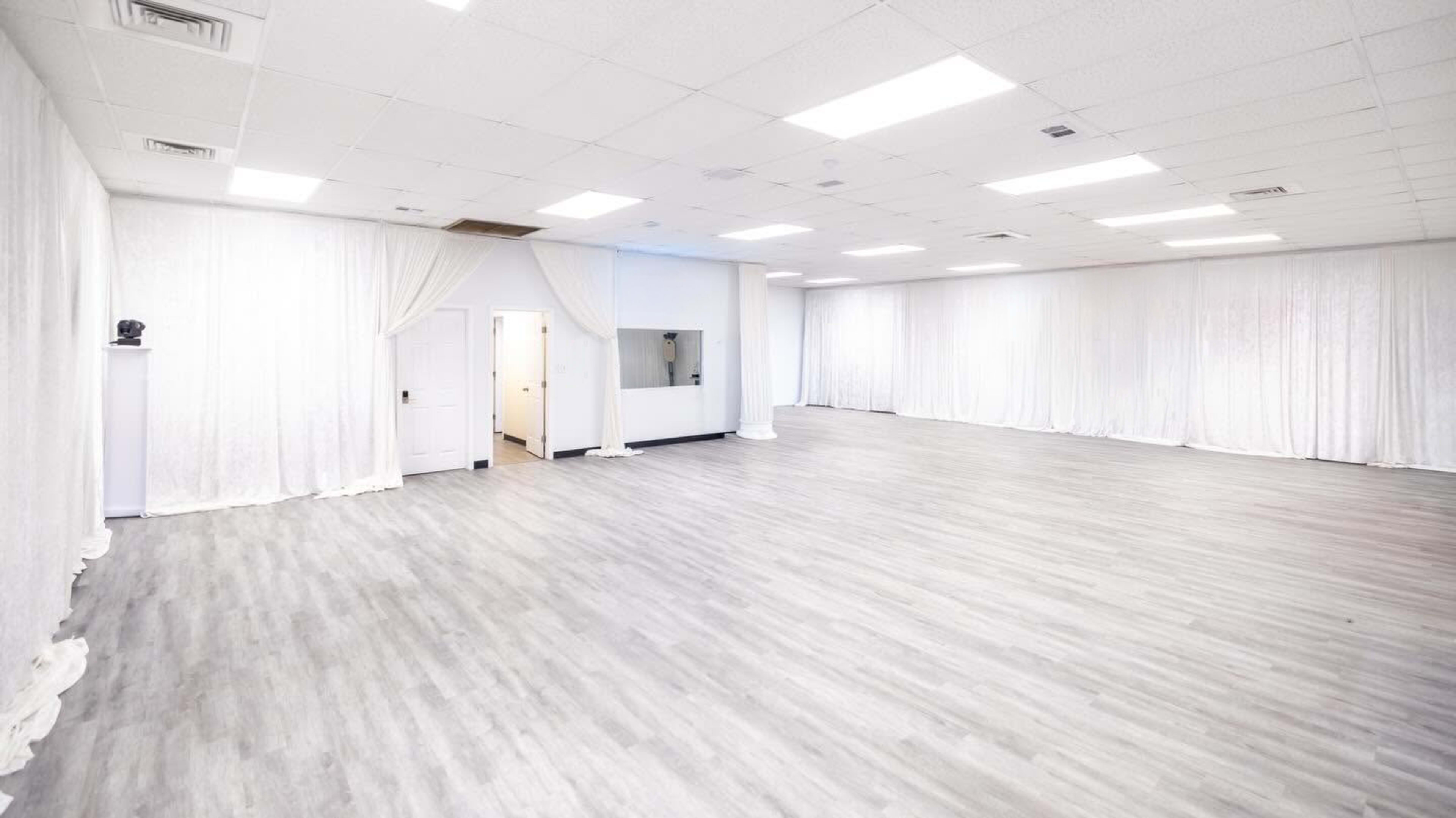 The image shows a spacious, empty room with light gray wood flooring and white drapes along the walls.