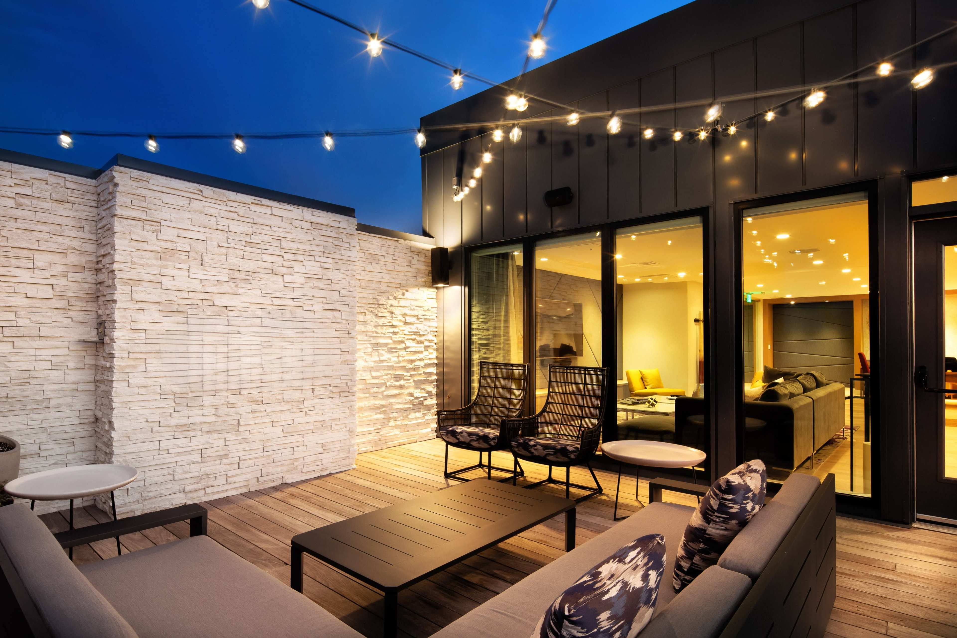 A modern rooftop patio features string lights overhead, with a sectional sofa, coffee table, and two chairs on a wooden deck, and glass doors leading into a well-lit interior space.