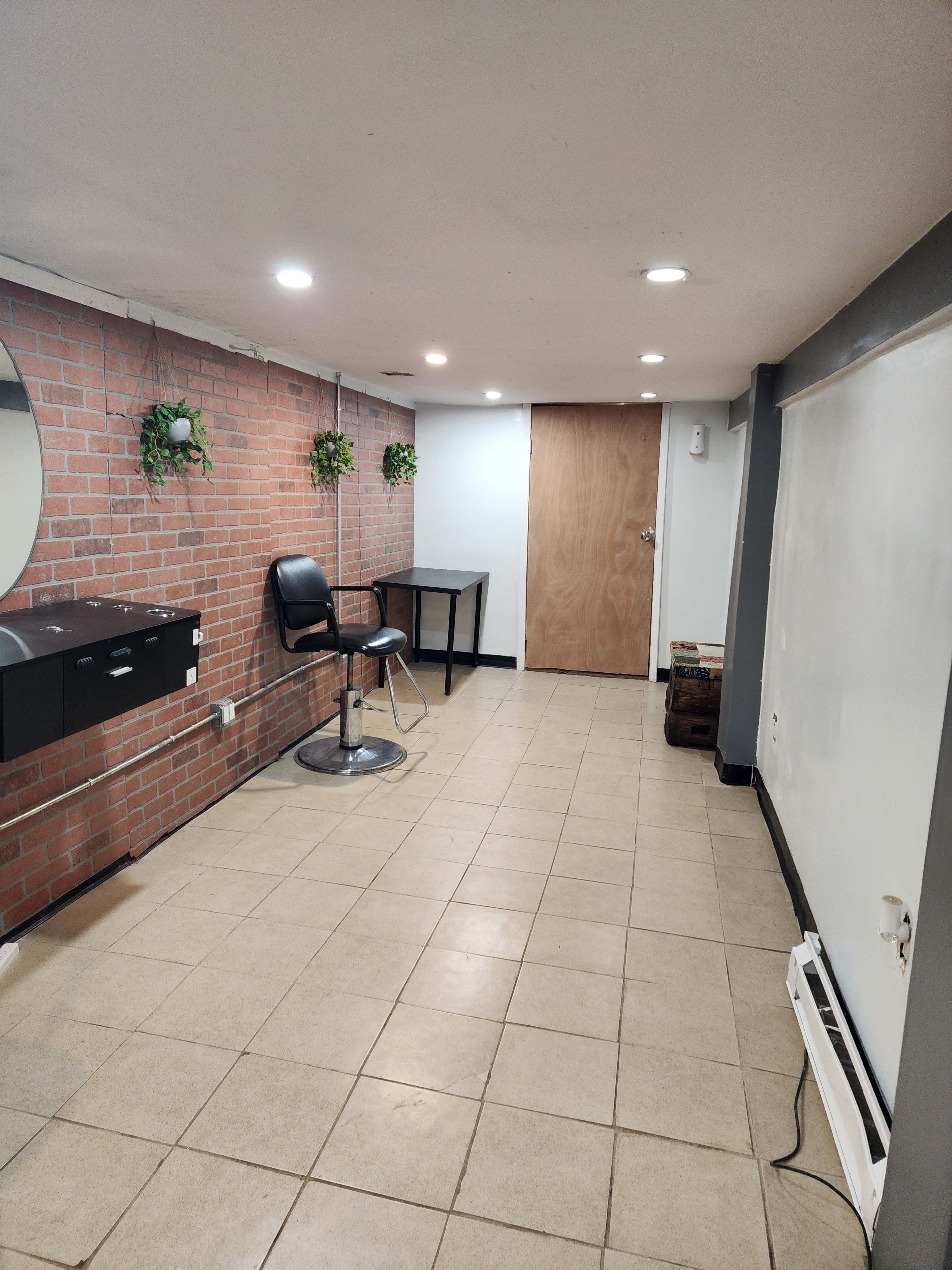 A clean, empty room with a brick wall, a large mirror, a black chair, a small table, and a door at one end.