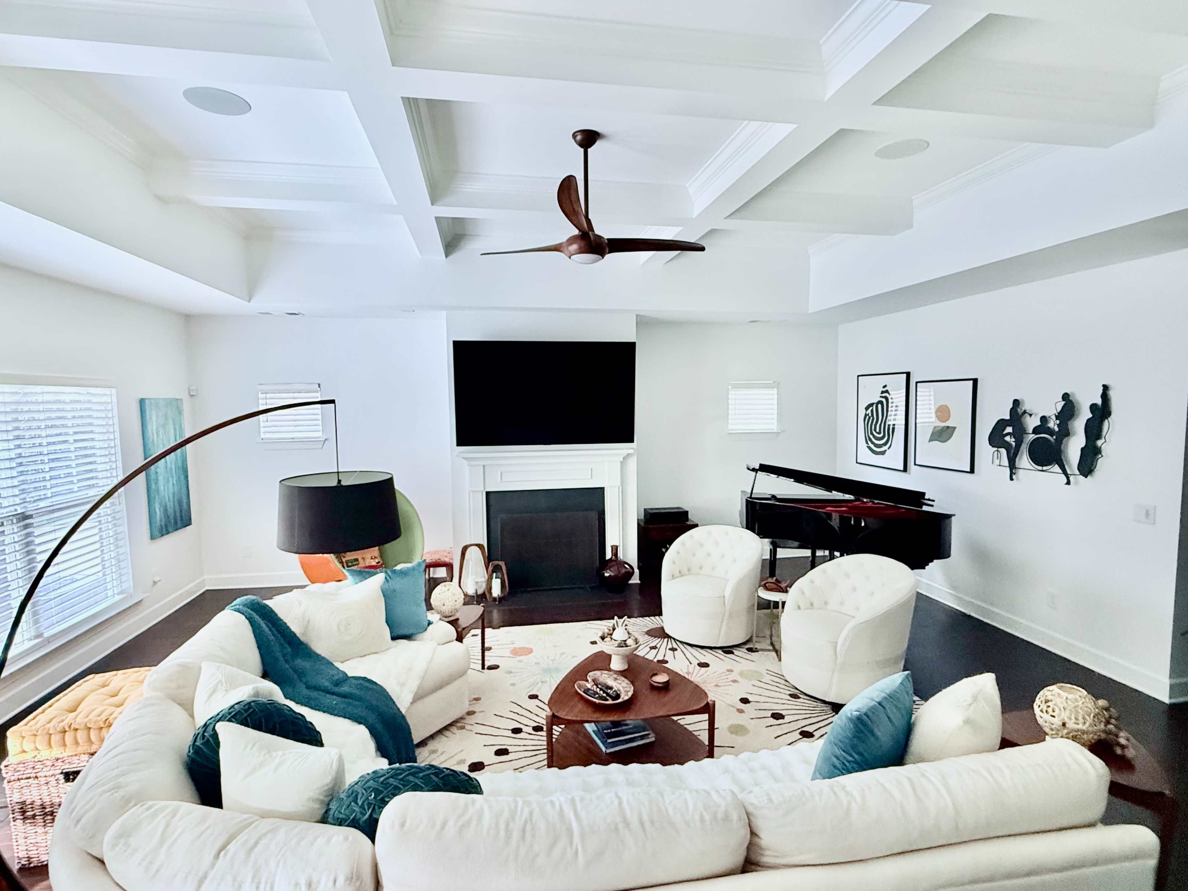 A spacious living room features a large sectional sofa, two armchairs, a piano, and a wall-mounted TV above a fireplace, all arranged around a round coffee table.