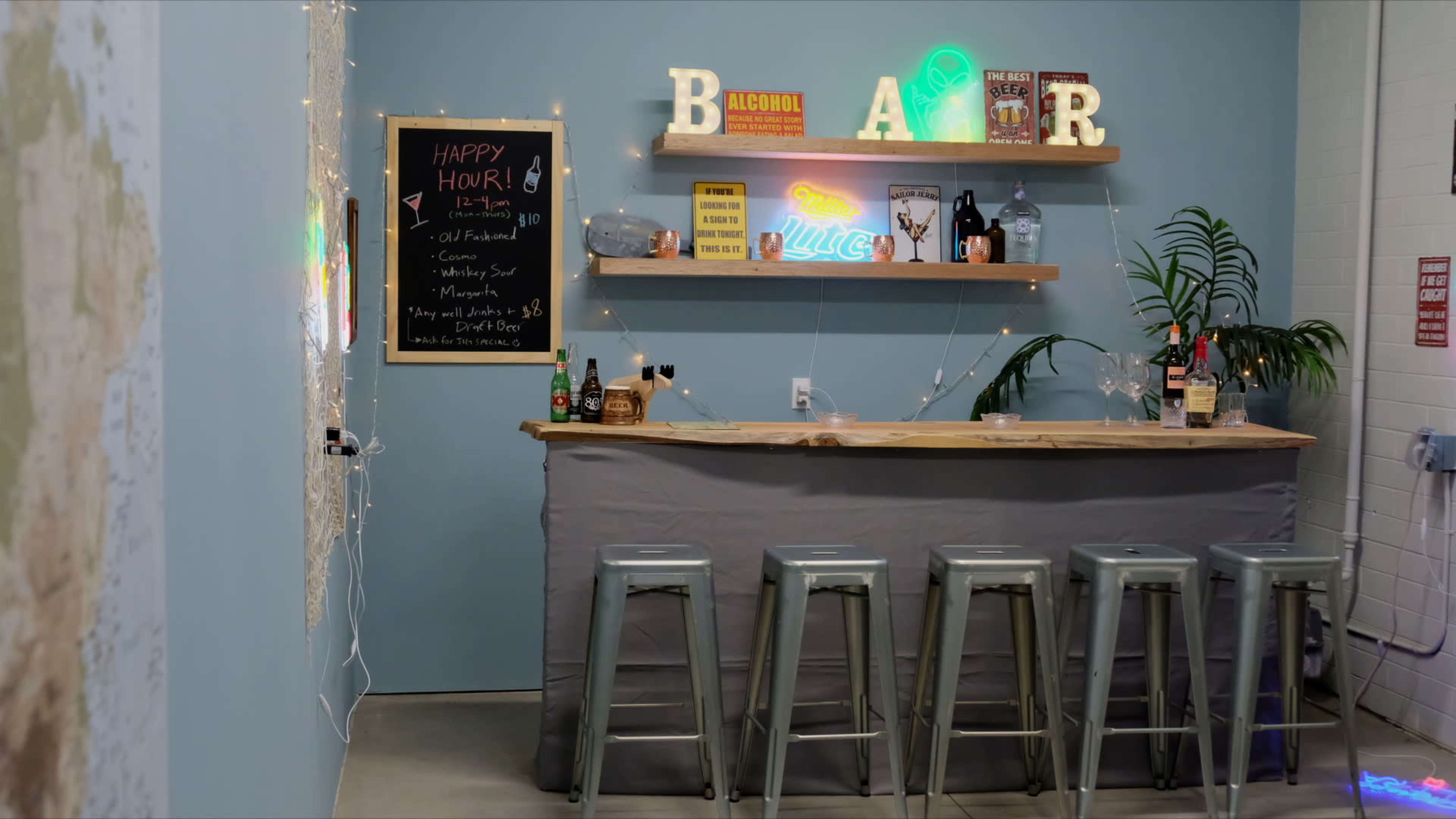 The image shows a small bar area with a wooden countertop, several metal stools, and neon signs against a blue wall, along with a chalkboard listing drink specials.