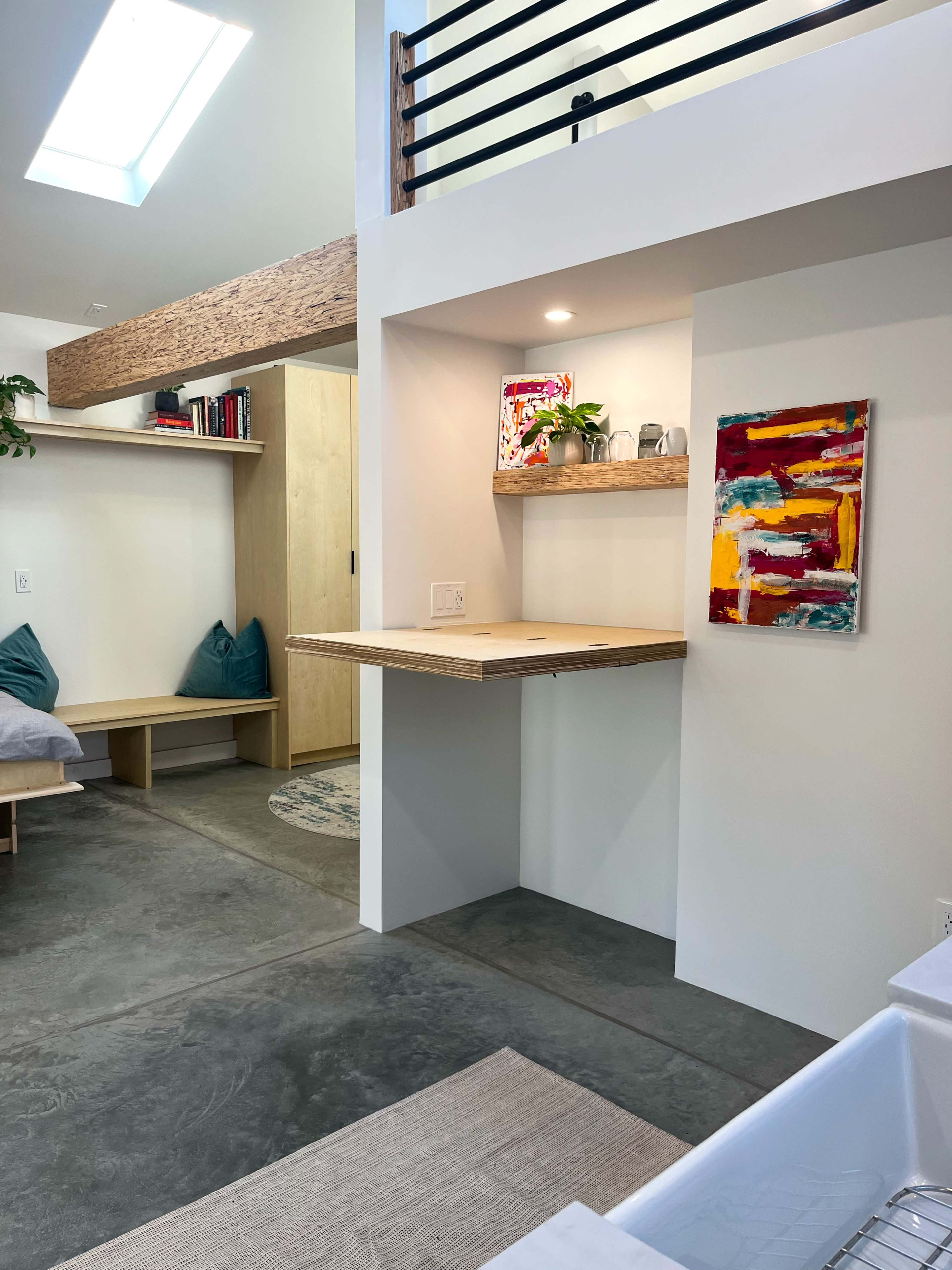 The image shows a modern interior space featuring a small kitchenette with a wooden shelf, a living area with a bed, and light-colored walls complemented by artistic wall decor.
