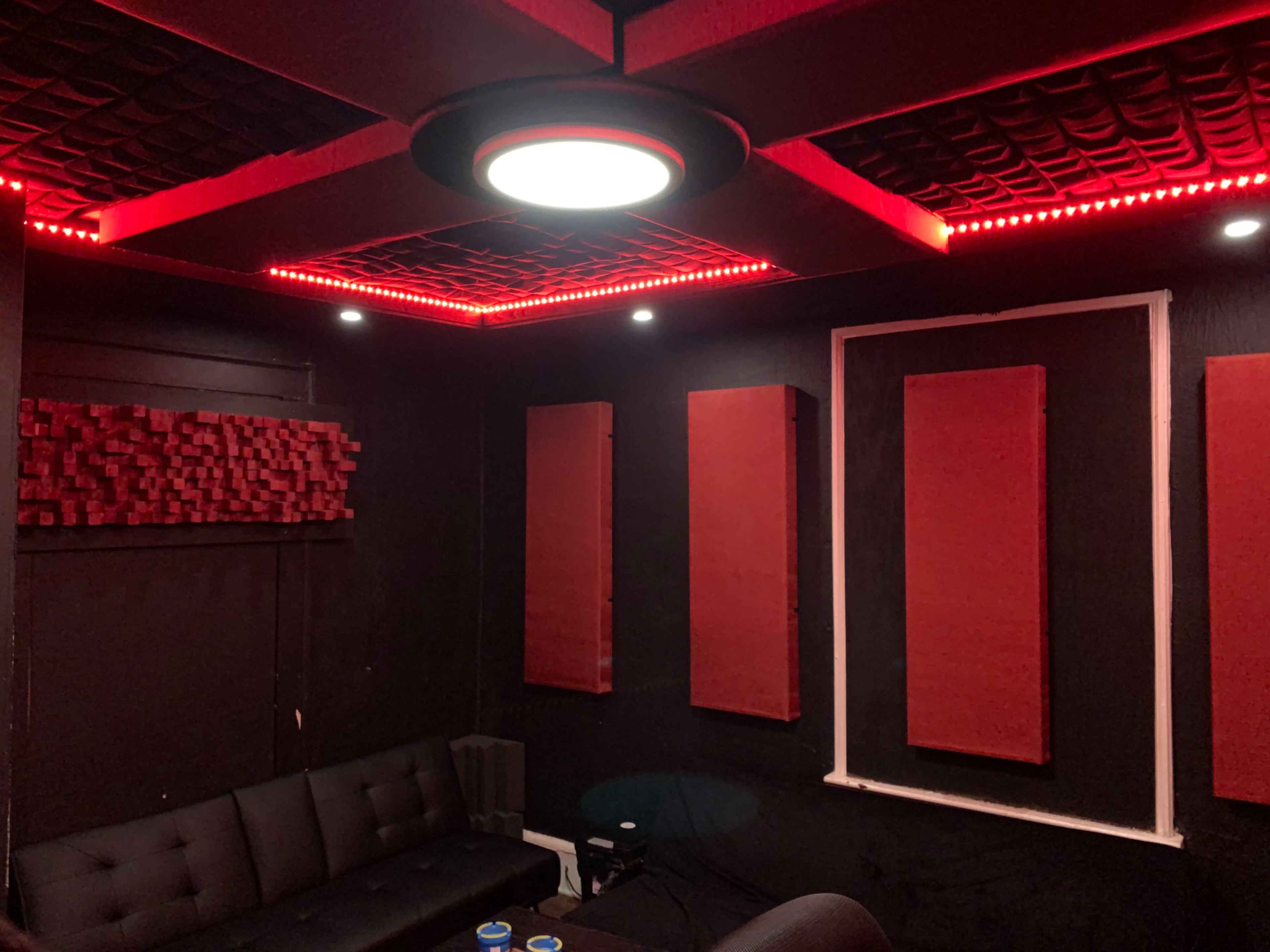 The image shows an indoor recording studio featuring black walls, a black sofa, red acoustic panels, and LED lights along the ceiling.