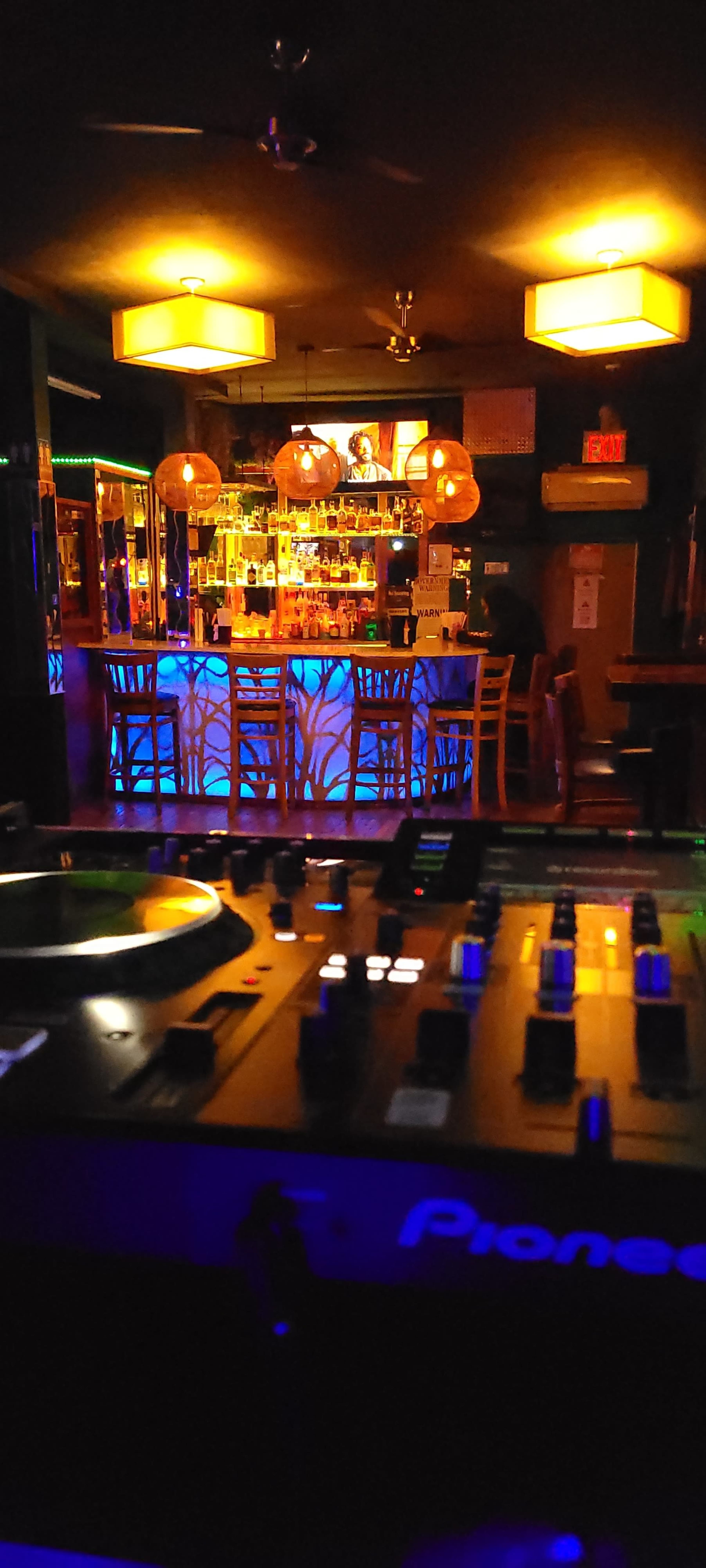 The image shows a DJ setup in the foreground with a vibrant bar illuminated by amber lighting in the background.