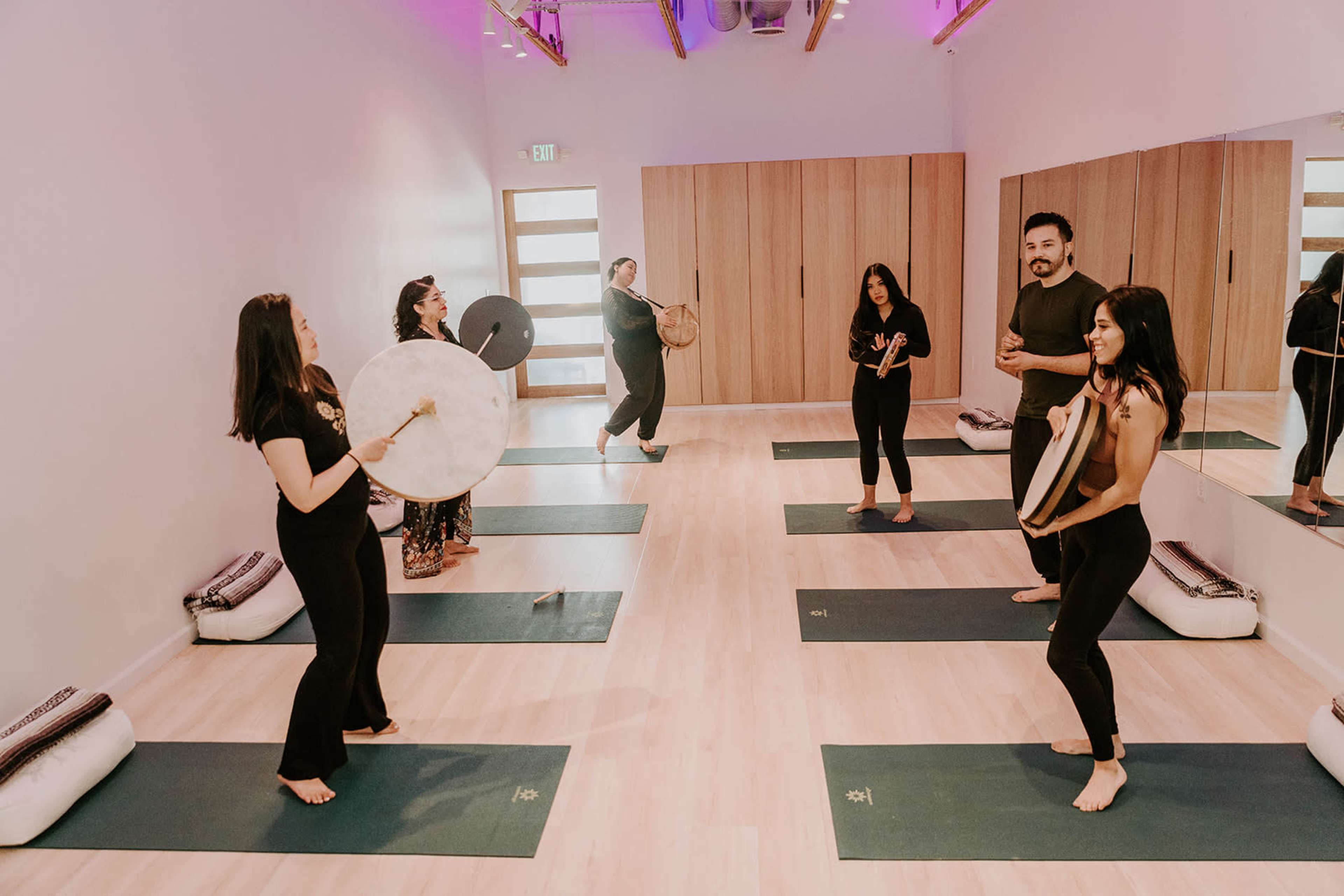 A group of six individuals engage in a yoga class, using various instruments, within a brightly lit studio with green mats and wooden accents.