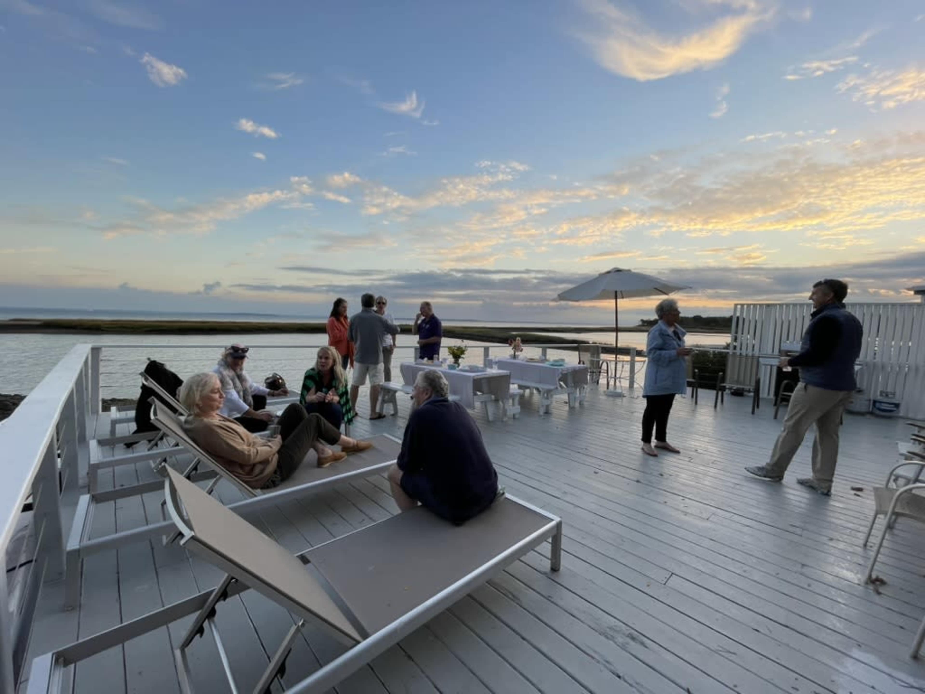 A group of people gathers on a wooden deck overlooking a body of water during sunset.
