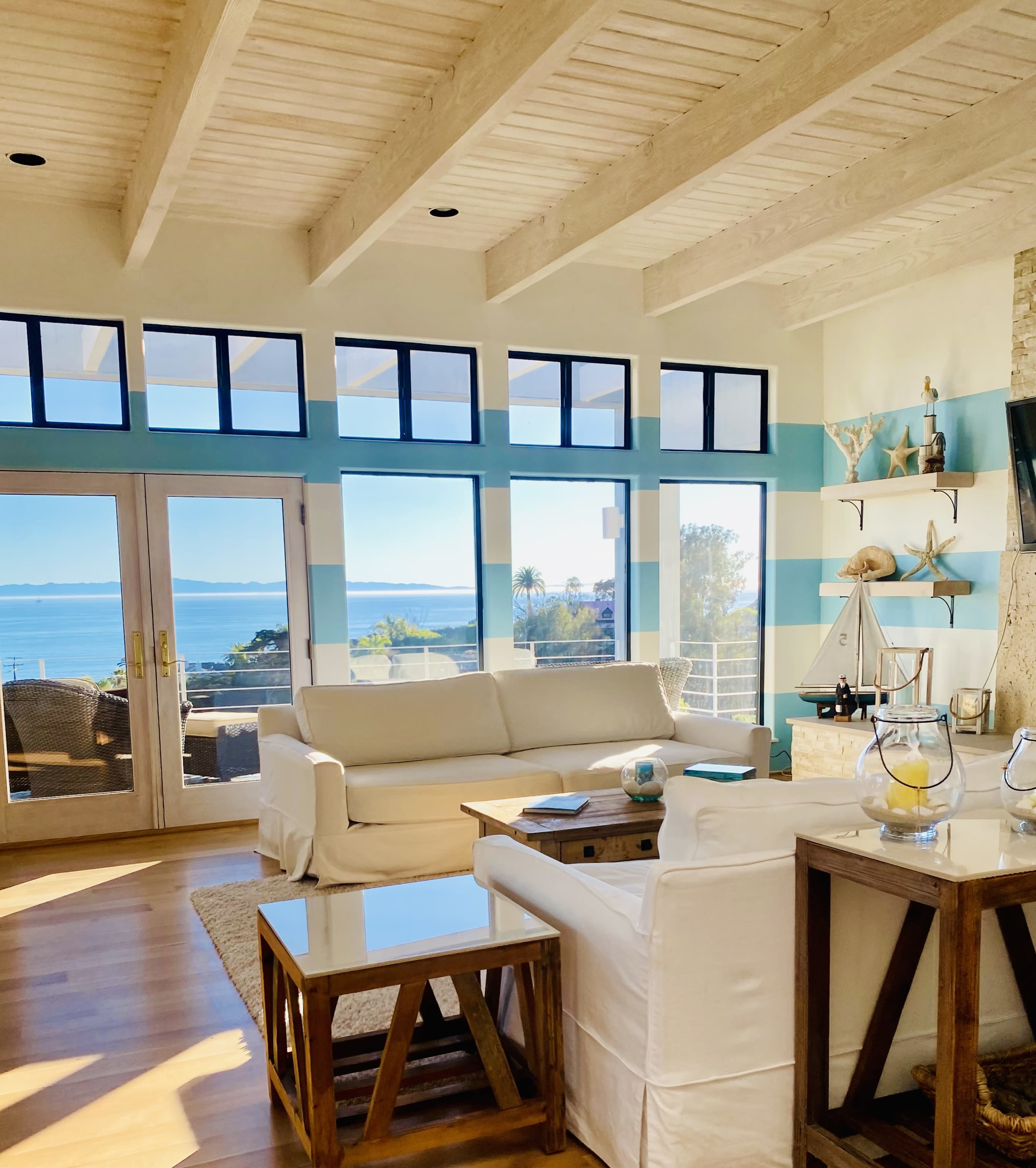A bright, modern living room features white furniture and large windows overlooking a scenic view of the ocean.