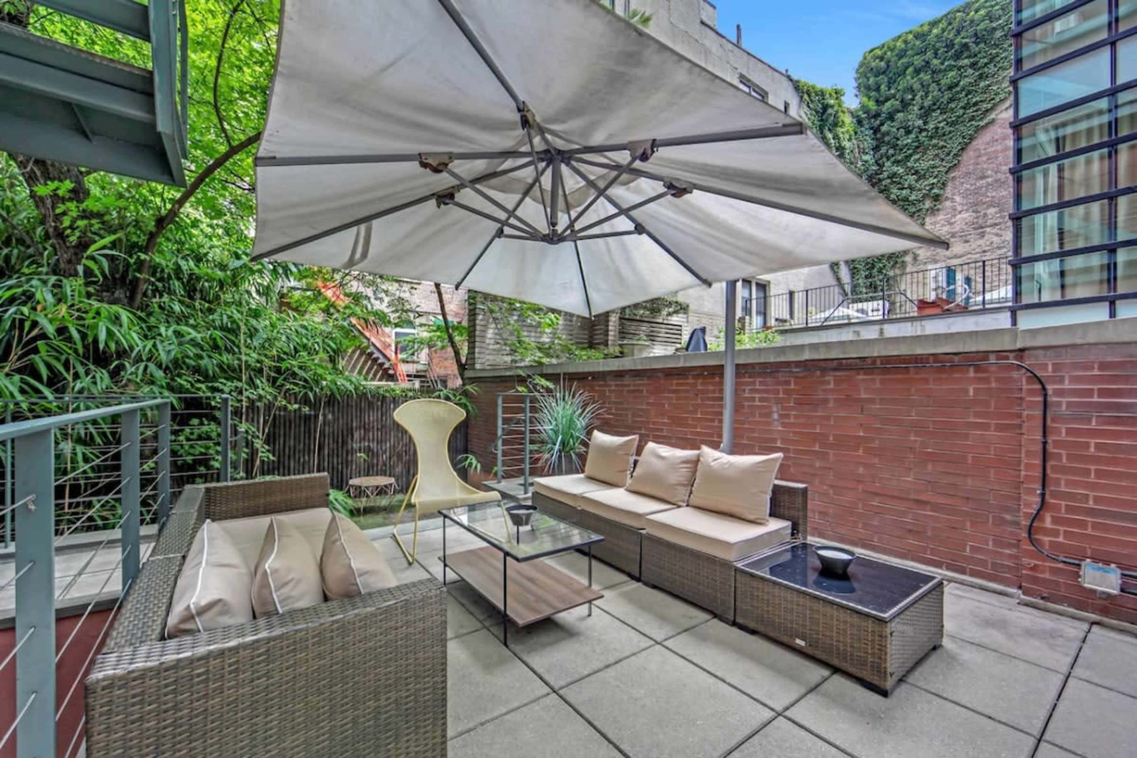 The image shows a cozy outdoor seating area with a large umbrella, a wicker sofa, and a glass coffee table against a backdrop of green foliage and brick walls.
