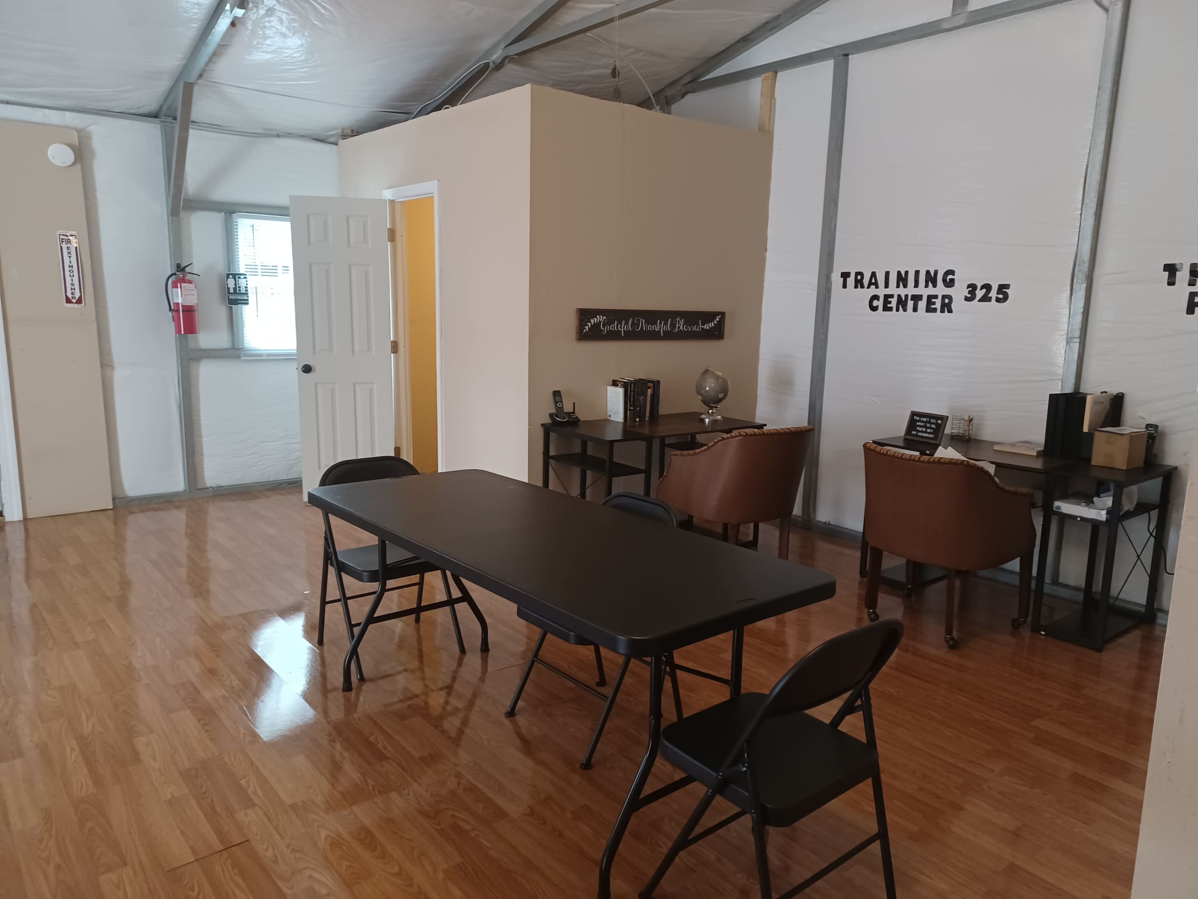 The image shows an interior of a training center featuring a table with chairs, a door leading to another room, and two chairs beside a desk.