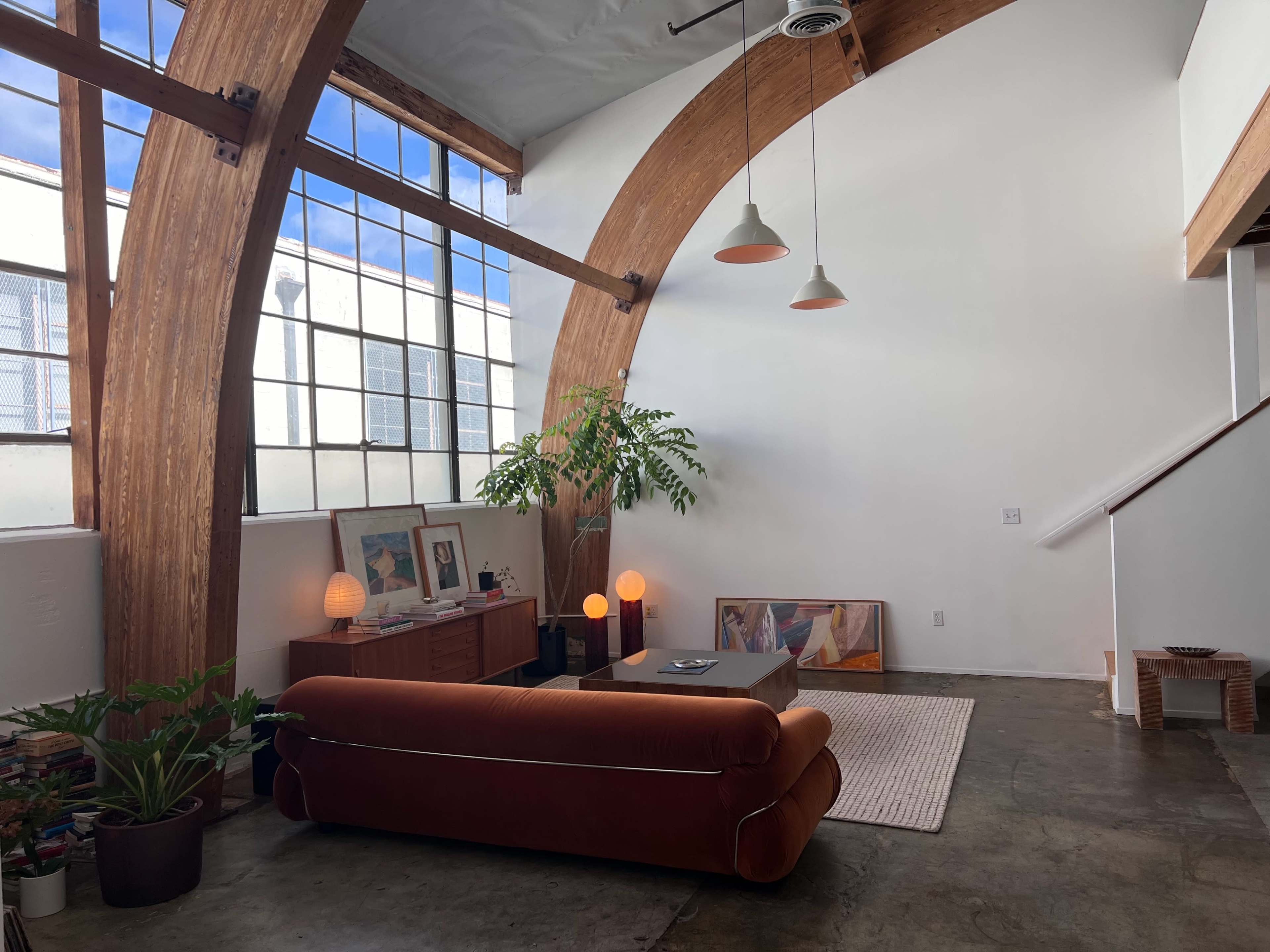 A spacious interior with wooden arches, a large window, a brown couch, a coffee table, and plants, all illuminated by pendant lights.