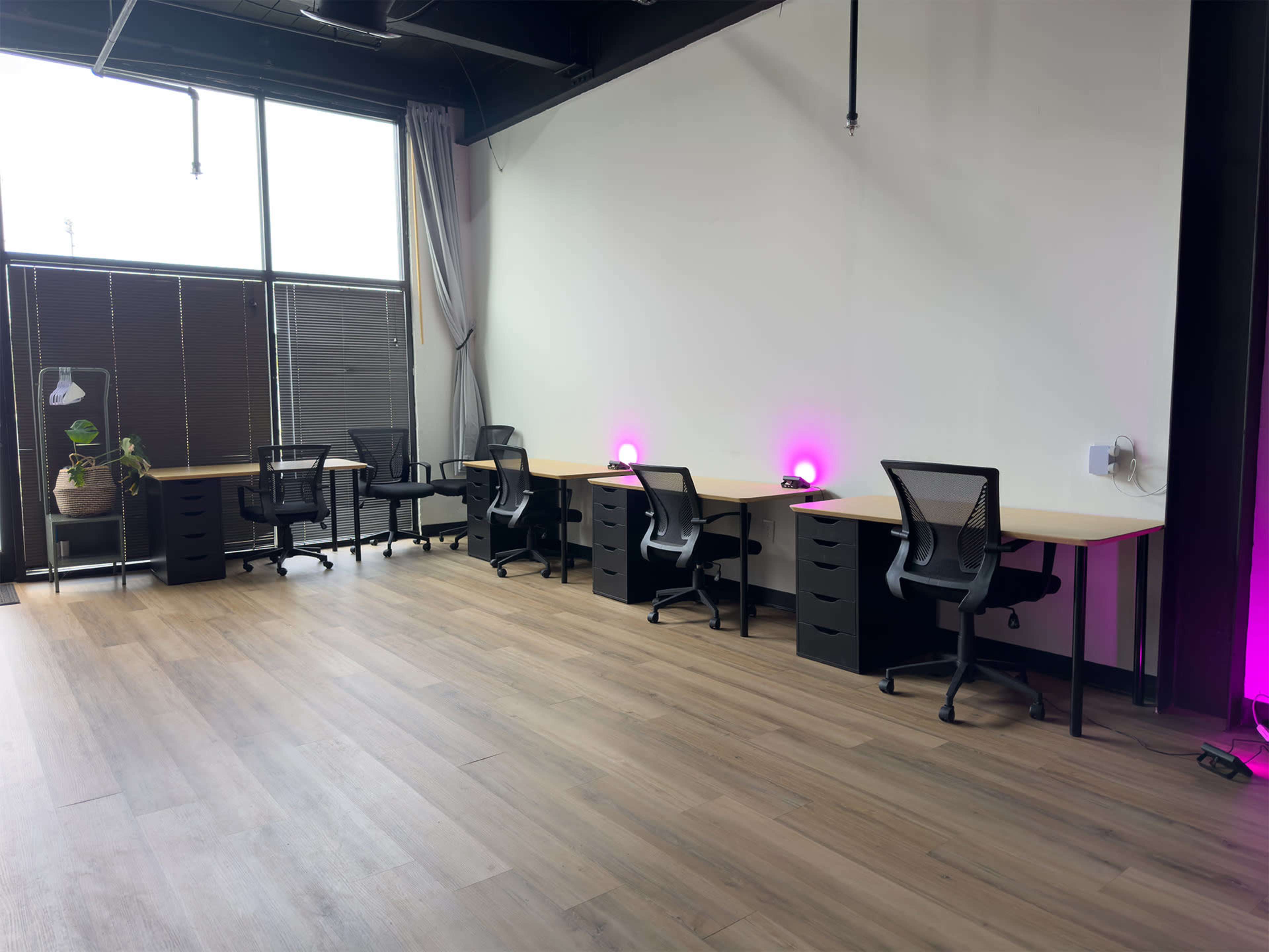 A coworking space with several desks and ergonomic chairs, illuminated by purple lights, along with large windows letting in natural light.