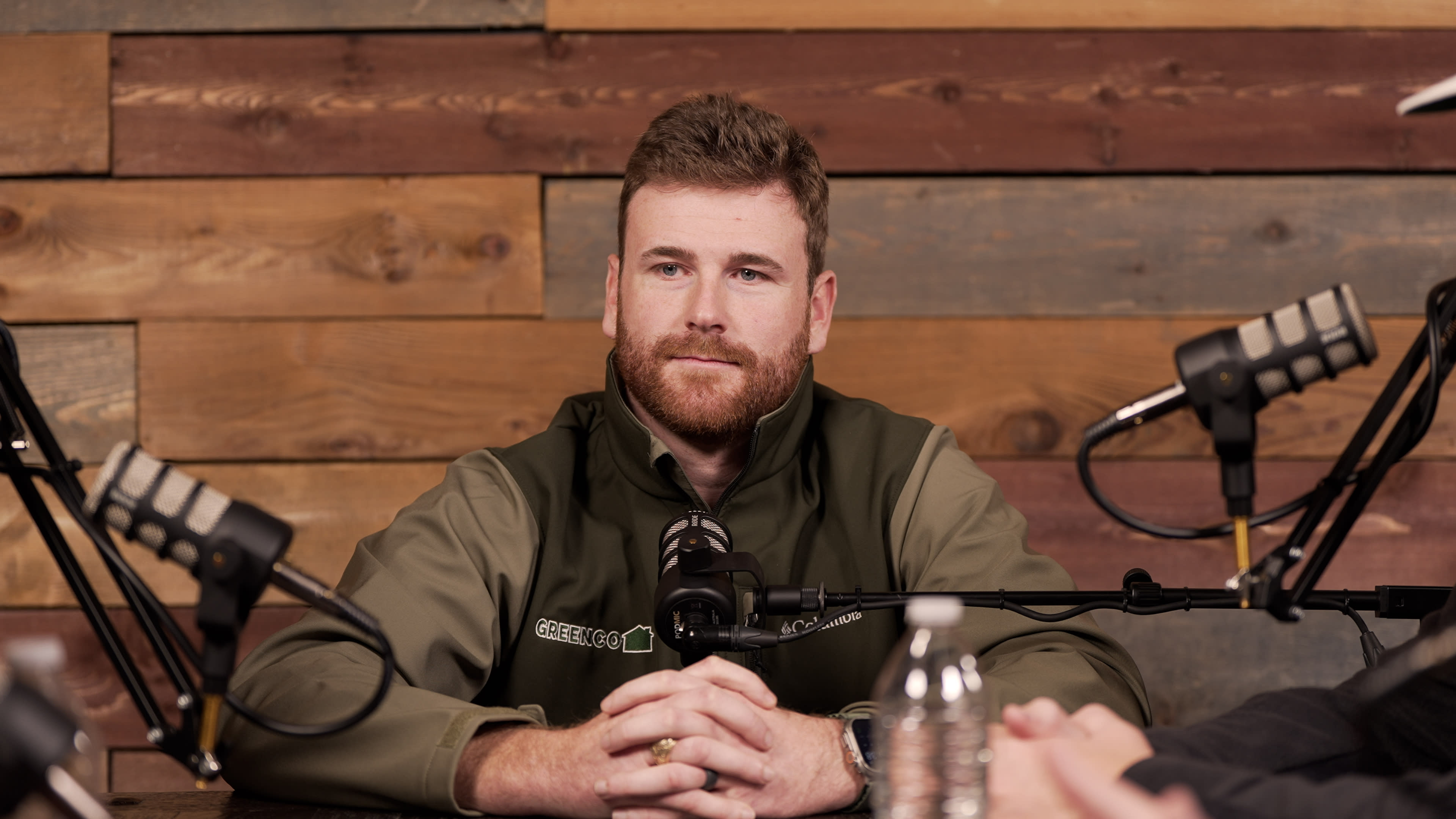 A man with a beard sits at a table in front of microphones, surrounded by wooden wall panels.