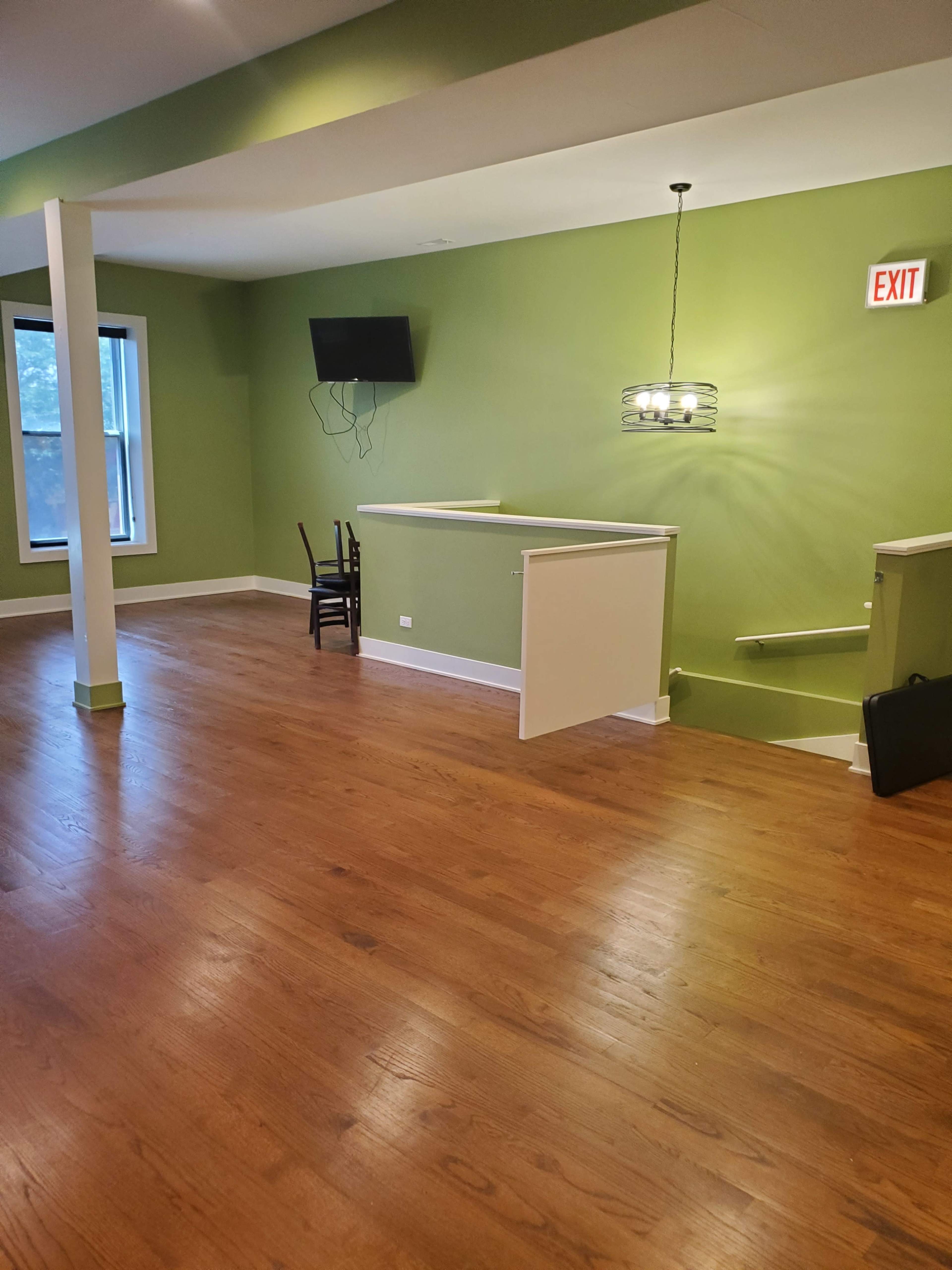 The image shows a spacious, empty room with green walls, hardwood flooring, a wall-mounted TV, a chandelier, and an exit sign.