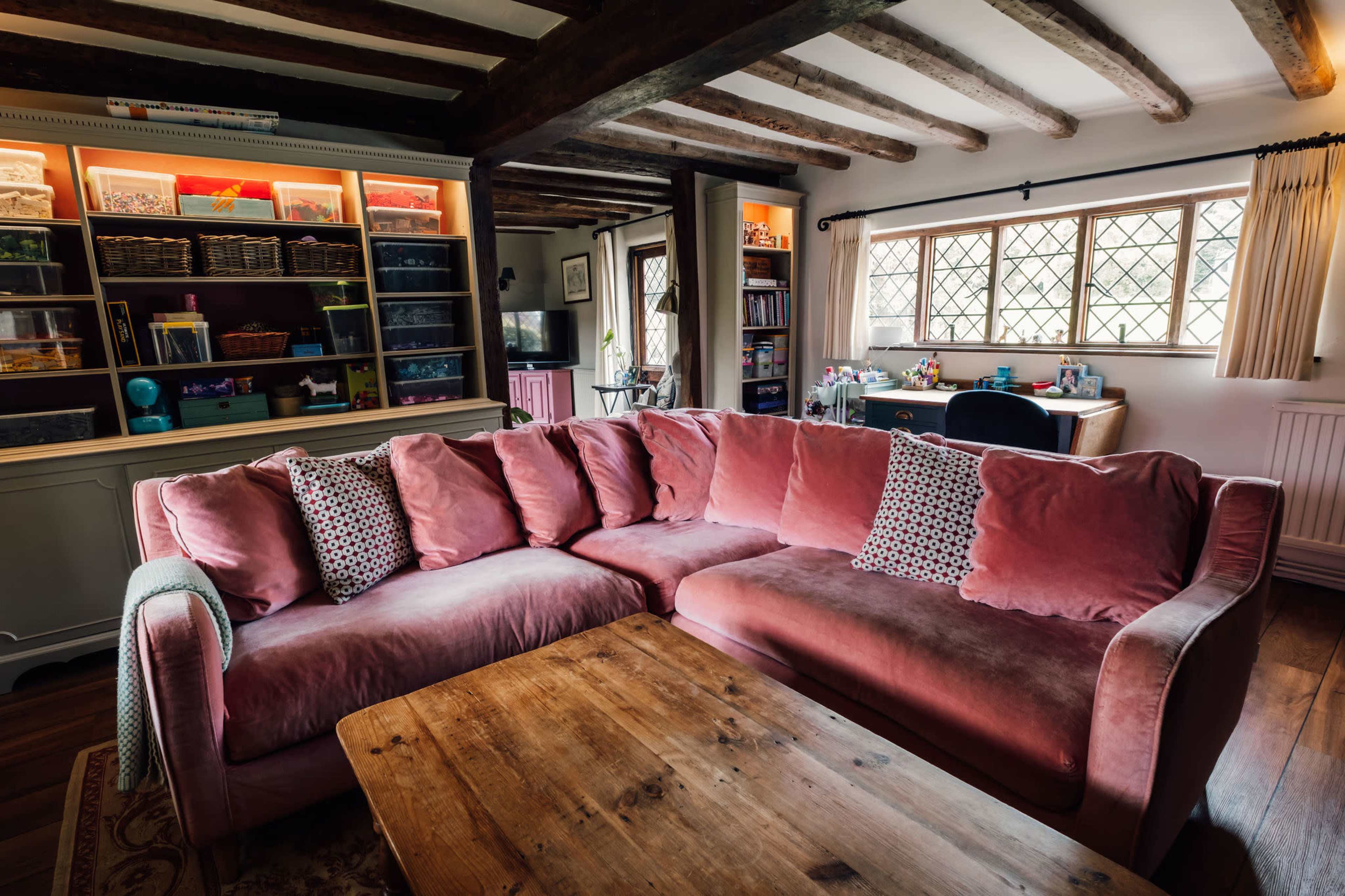 The image shows a cozy living room with a pink sectional sofa, a wooden coffee table, and shelves filled with colorful storage boxes in the background.