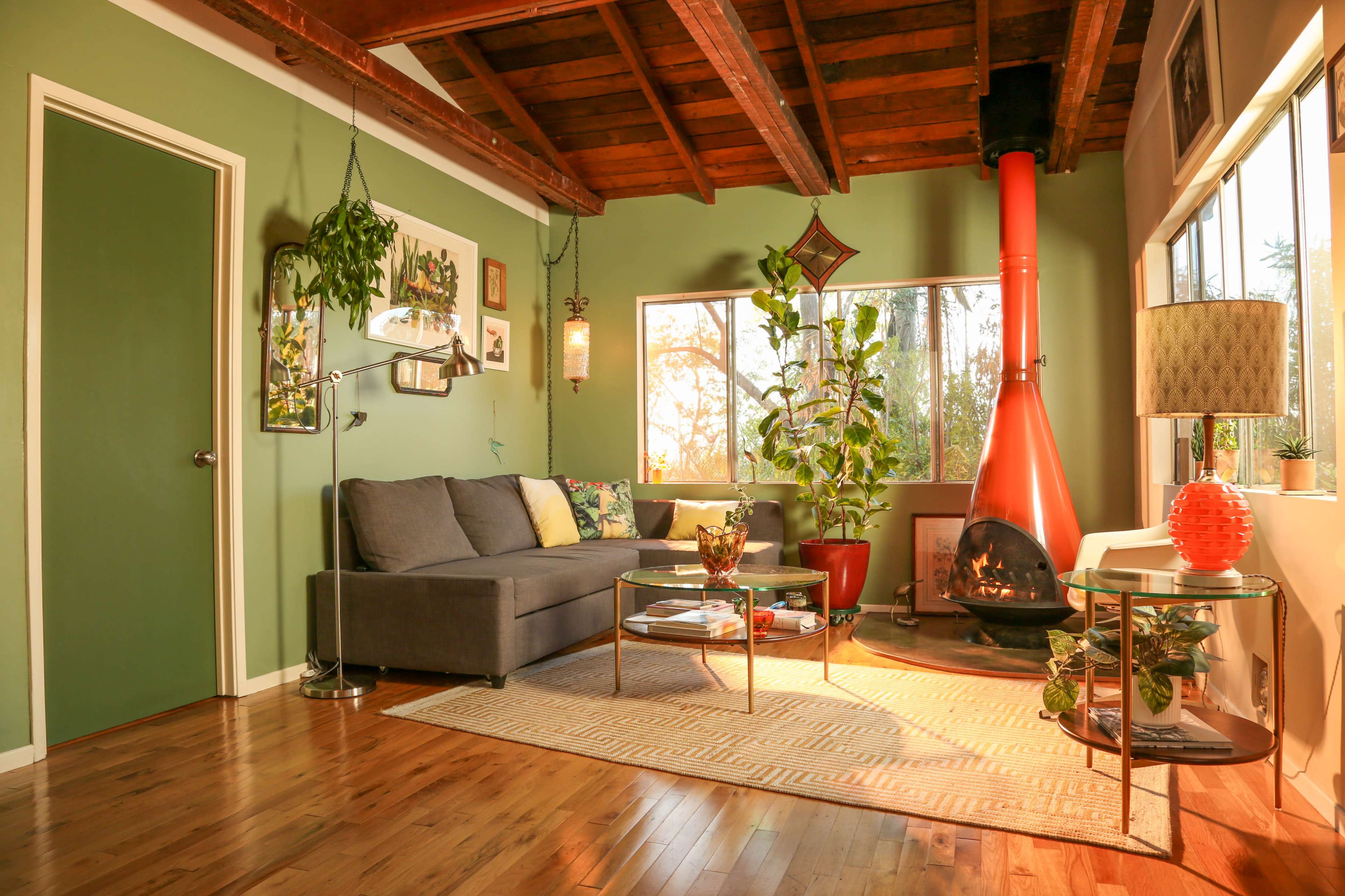 The image depicts a cozy living room featuring a gray sofa, a circular coffee table, a wood-burning stove with a red chimney, and several plants positioned near the windows.