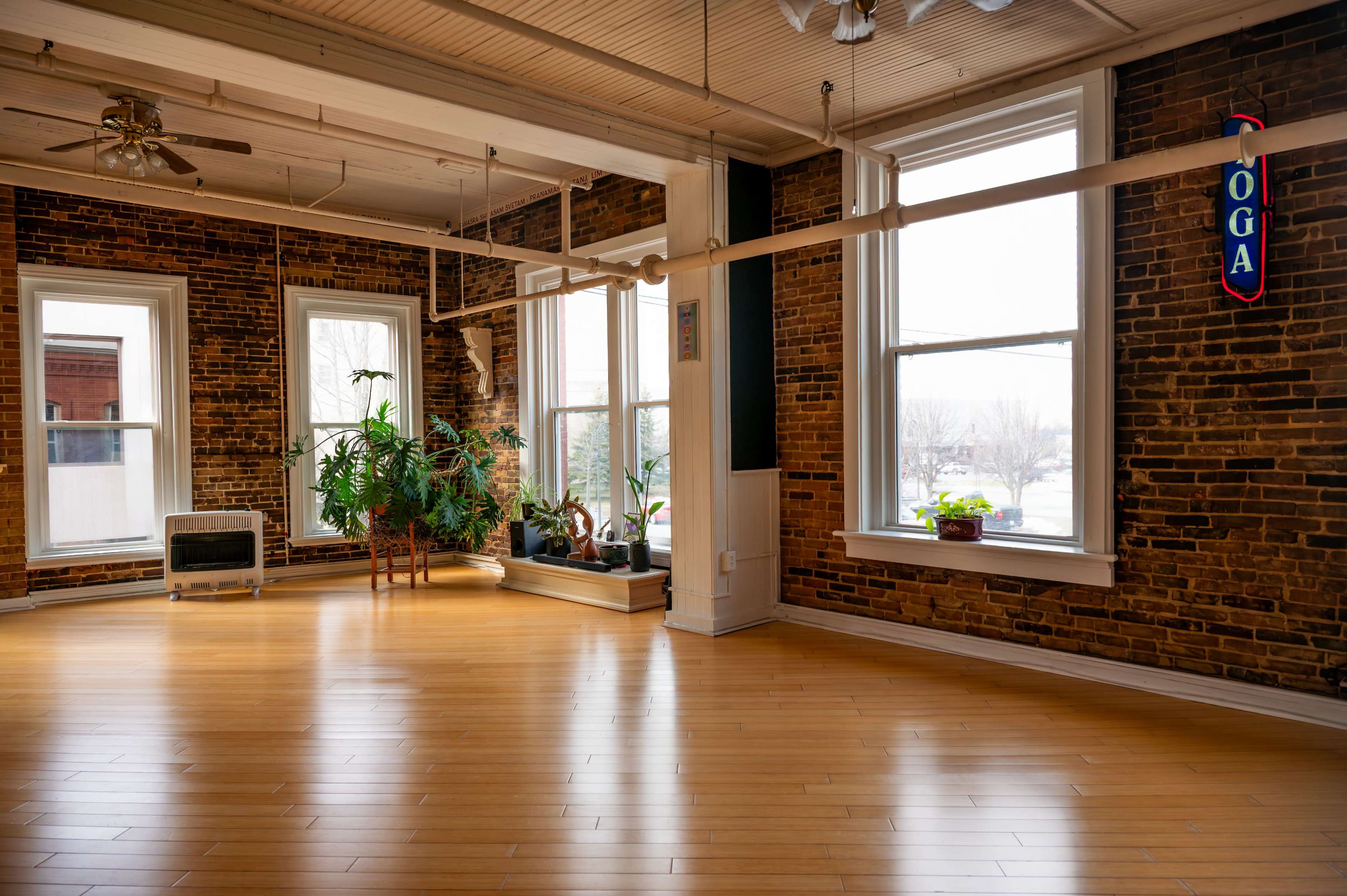 The image shows a spacious yoga studio with exposed brick walls, large windows, and wooden flooring.