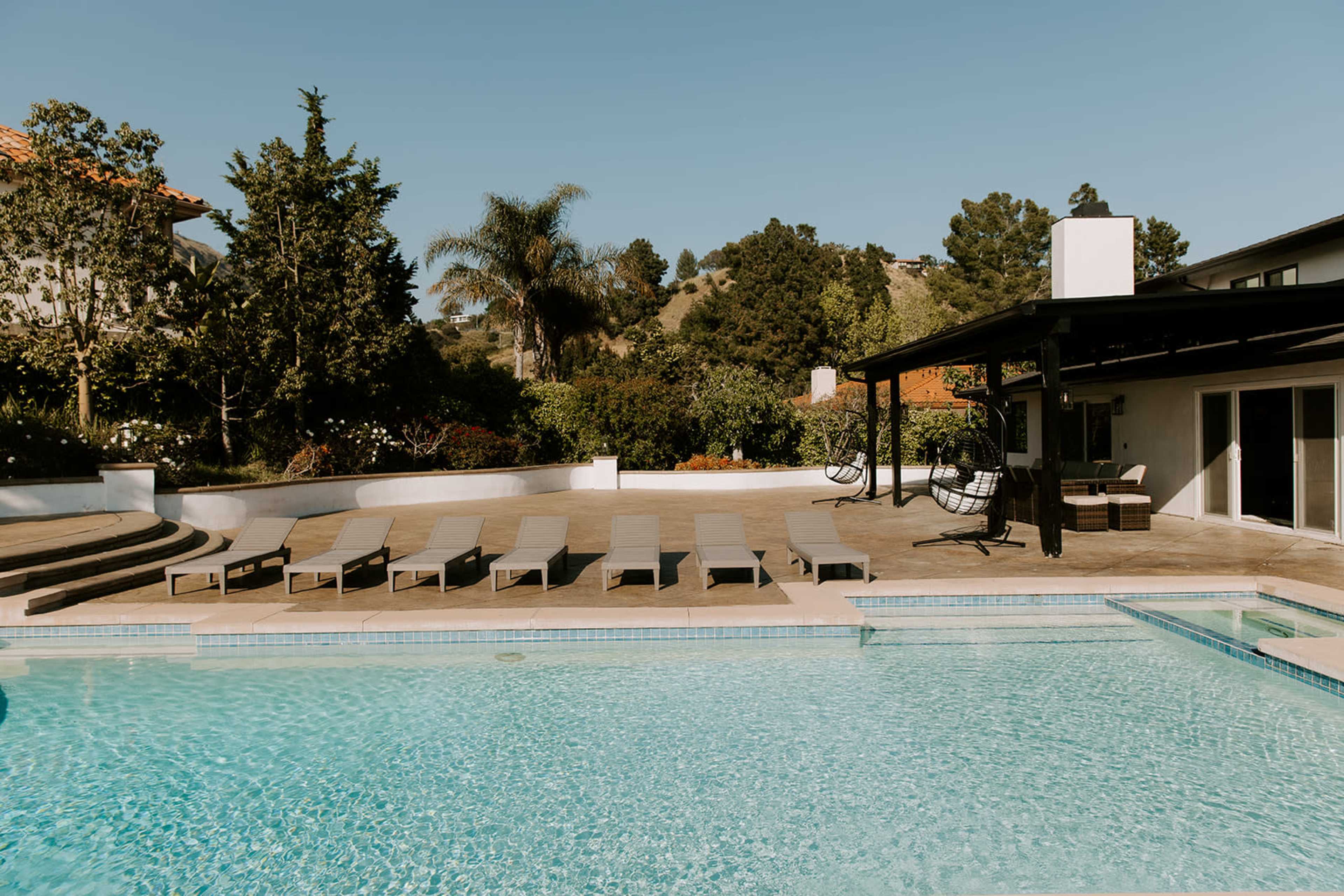 A clear swimming pool is surrounded by lounge chairs and a shaded seating area, with trees and hills in the background.