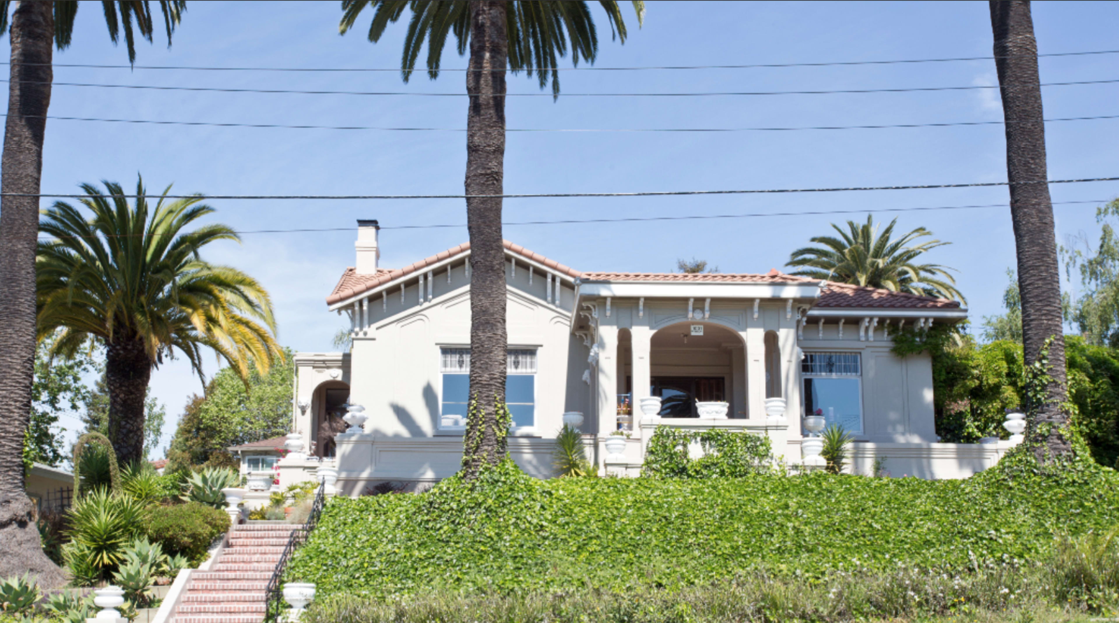 A large, two-story house is framed by palm trees and surrounded by lush greenery, with a brick pathway leading to the entrance.